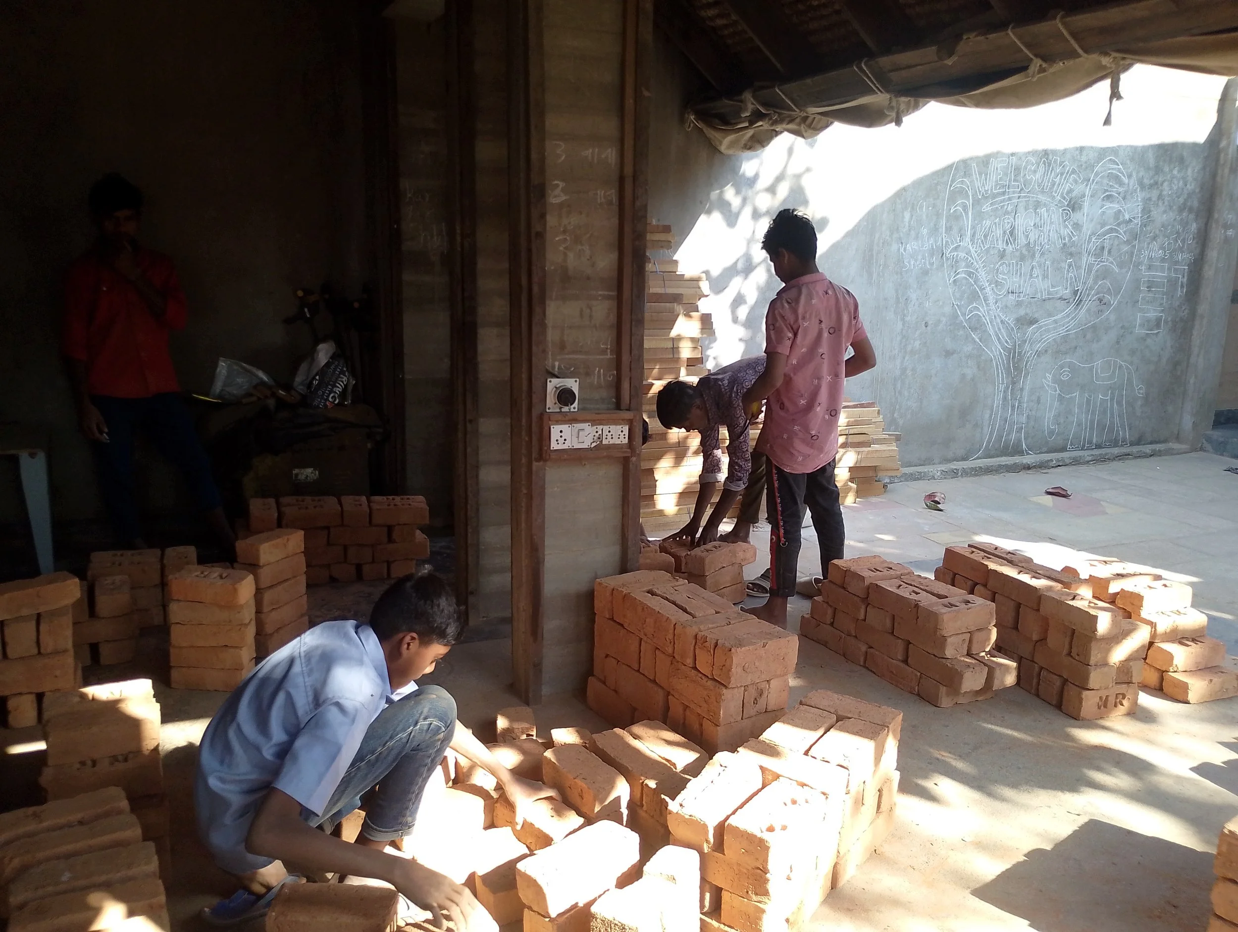 Young boys are building brick structures outdoors near a wall with a chalk drawing of a tree and an elephant. One boy is kneeling down, arranging bricks, while others are stacking bricks or working on the structure.