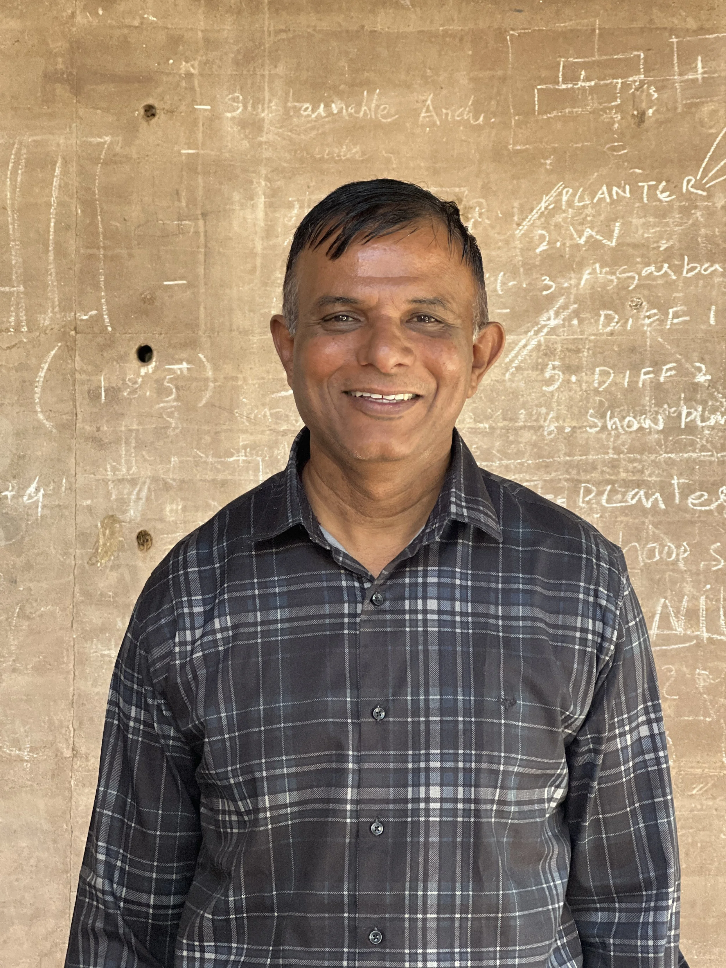 A smiling man in a black and gray plaid shirt standing in front of a beige wall with handwritten notes and diagrams.