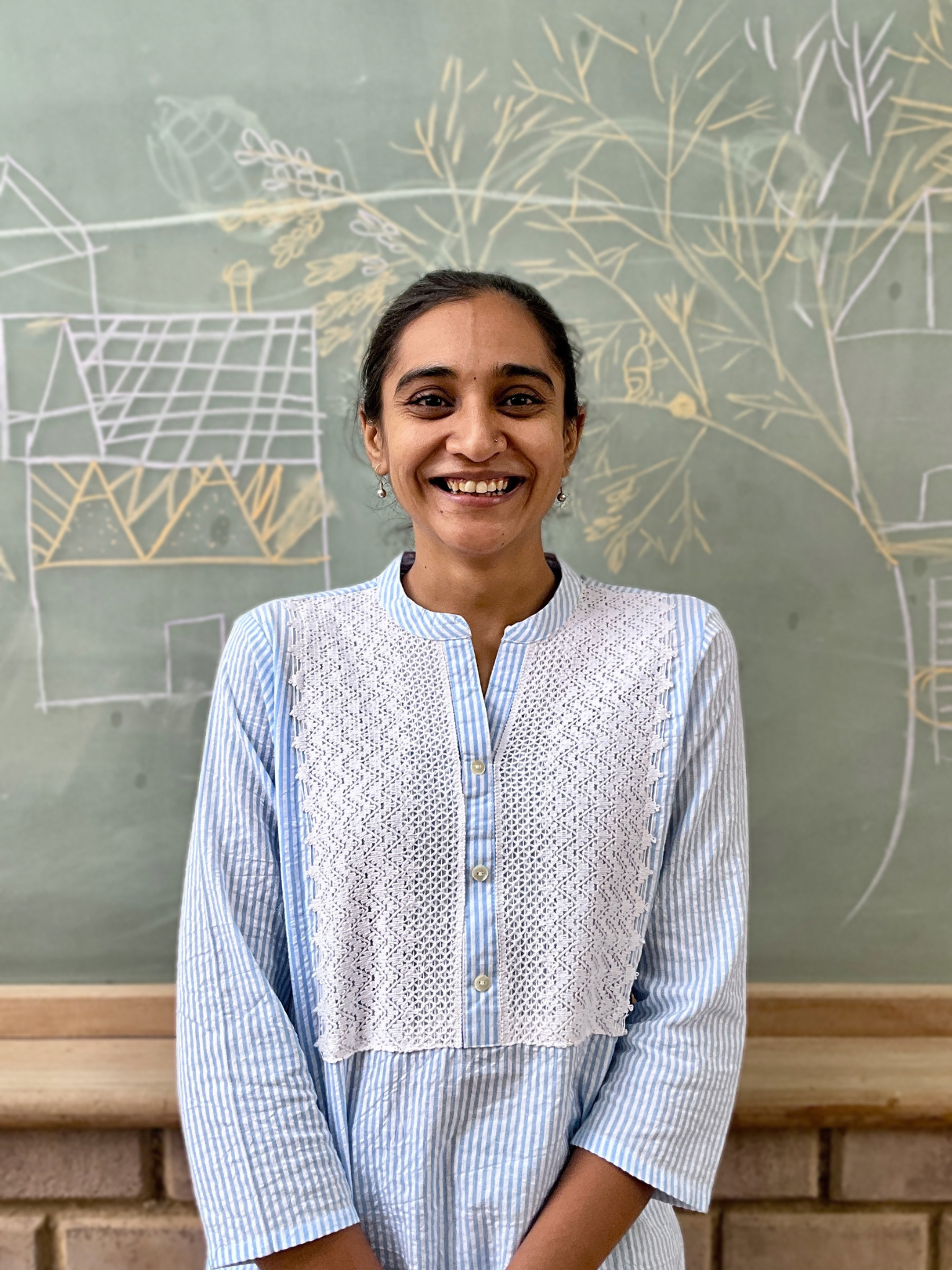 Woman smiling in front of a chalkboard with white and yellow chalk drawings of plants and structures.