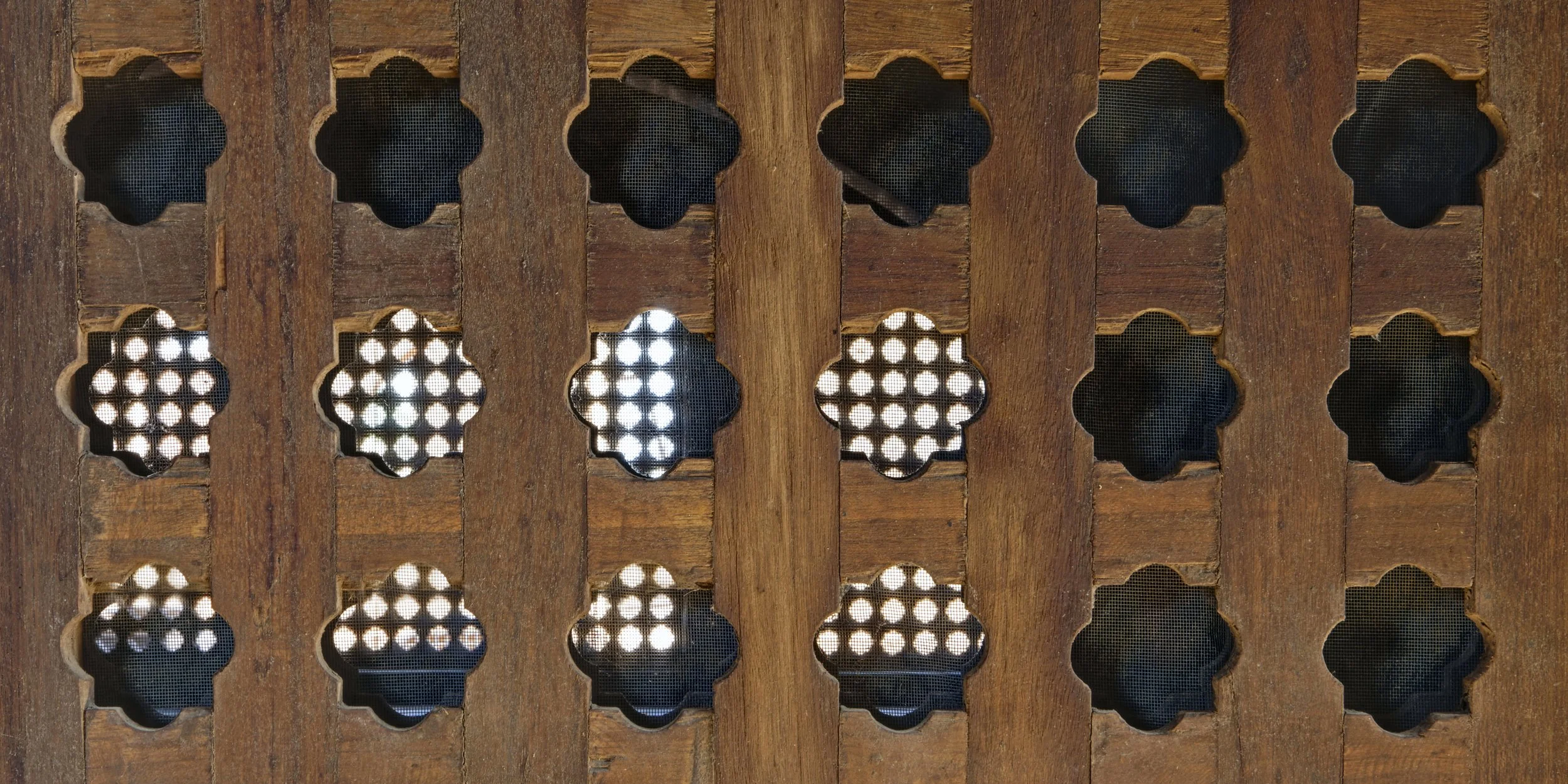 Wooden lattice with decorative cutouts, revealing patterned and perforated metal underneath.