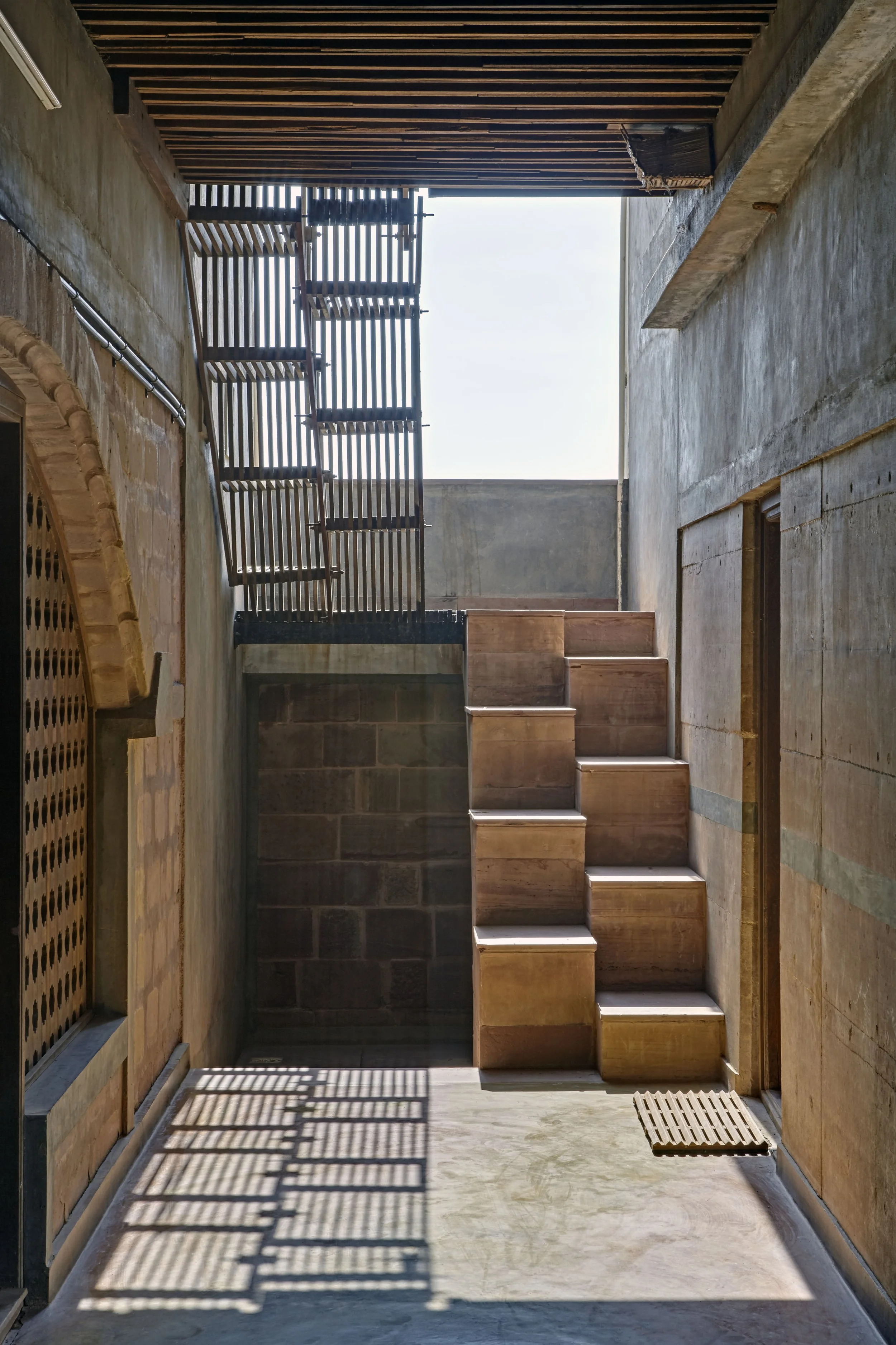 Interior view of a modern building with concrete and wooden stairs, wooden slat railing, exposed concrete walls, and shadows cast by the railing on the floor, with a blue sky visible through the open ceiling.