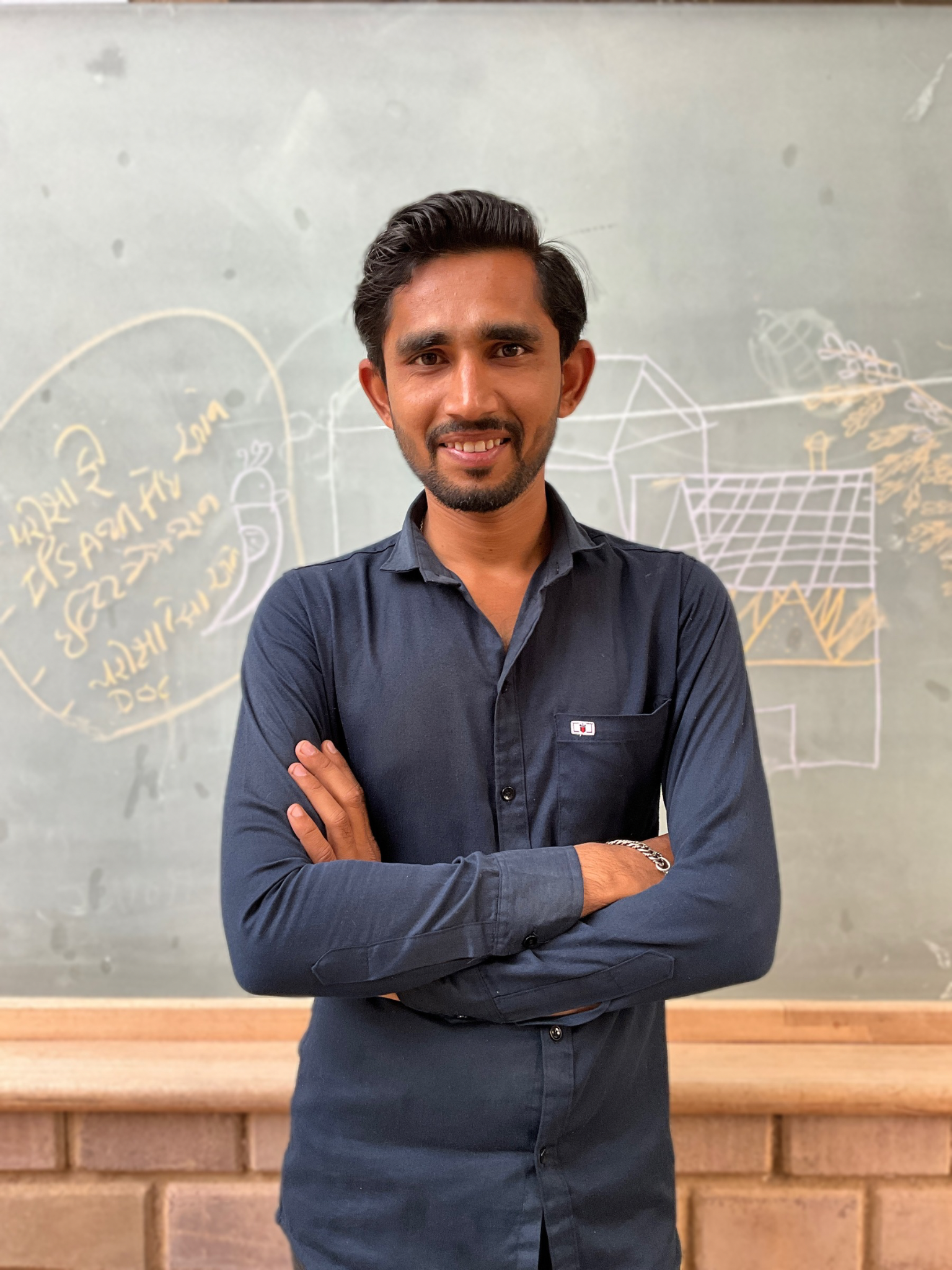 A man with dark hair and a beard smiling, standing in front of a wall with handwritten notes and diagrams.