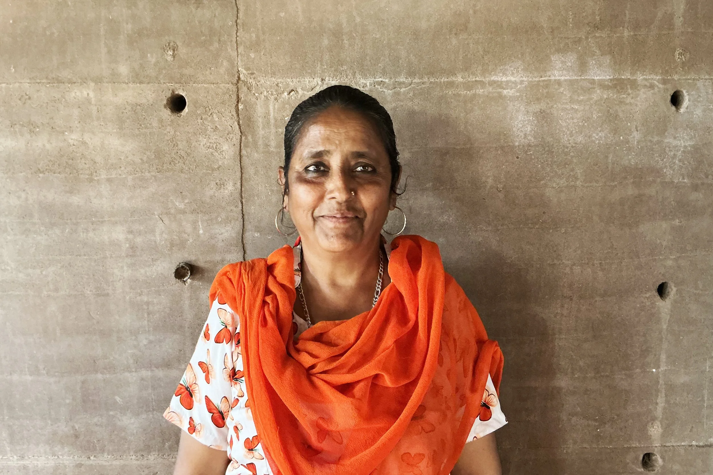 Woman with dark hair, wearing an orange scarf and butterfly-patterned blouse, standing against a concrete wall.