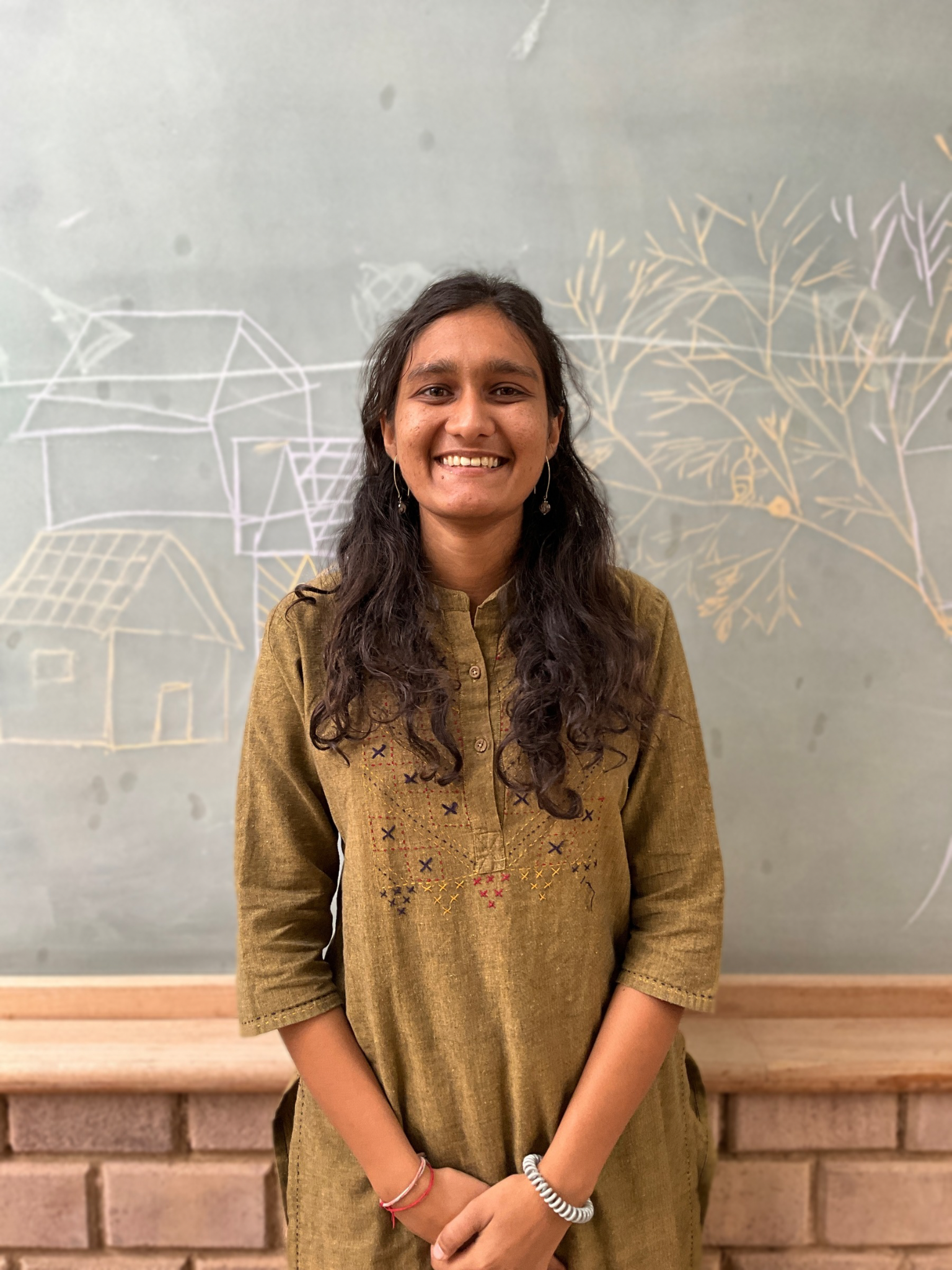 Young woman with long dark hair smiling, standing in front of a chalkboard with chalk sketches of buildings and trees.