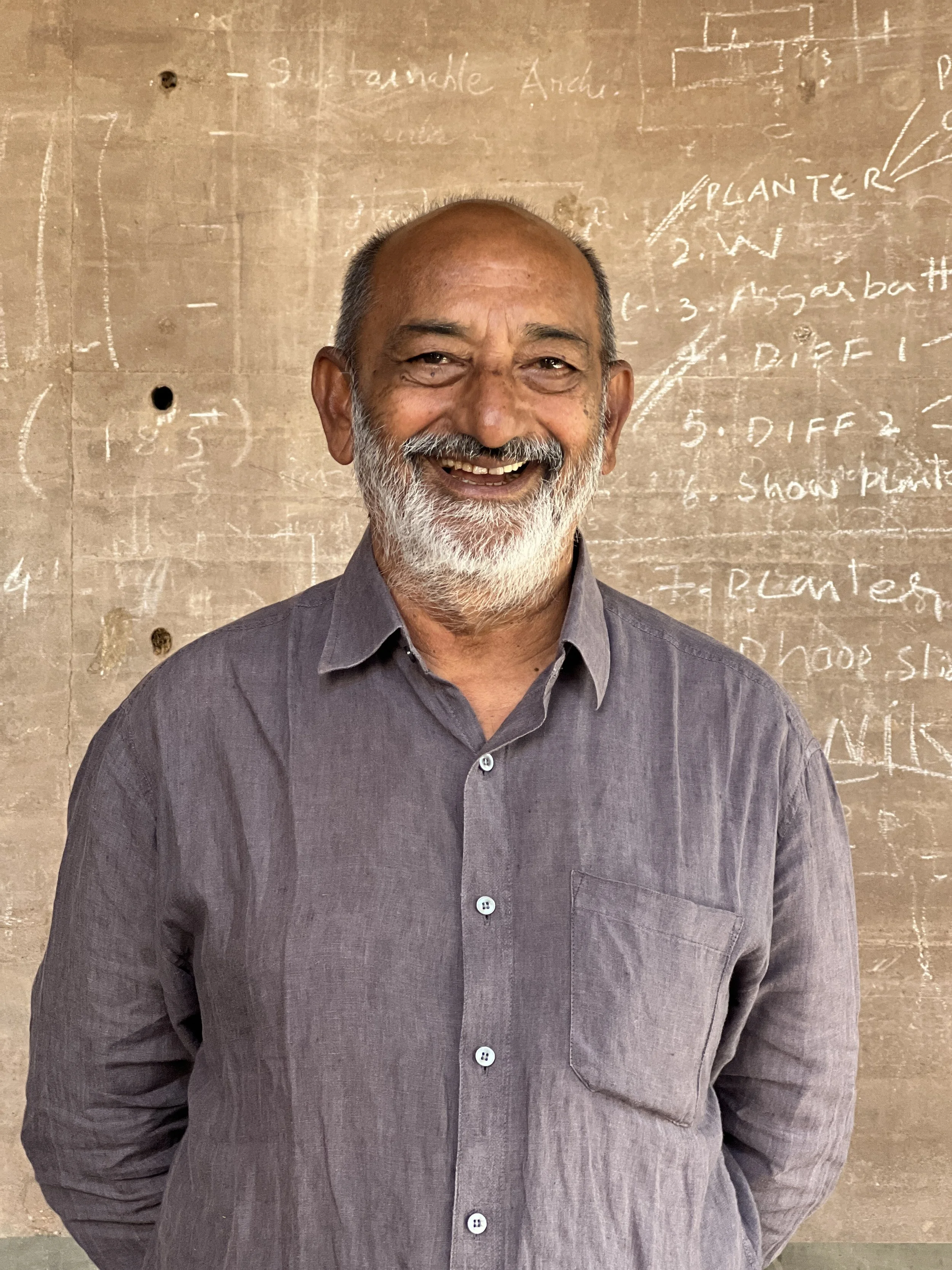 A smiling man with a beard and gray hair standing in front of a chalkboard with notes and diagrams written on it.