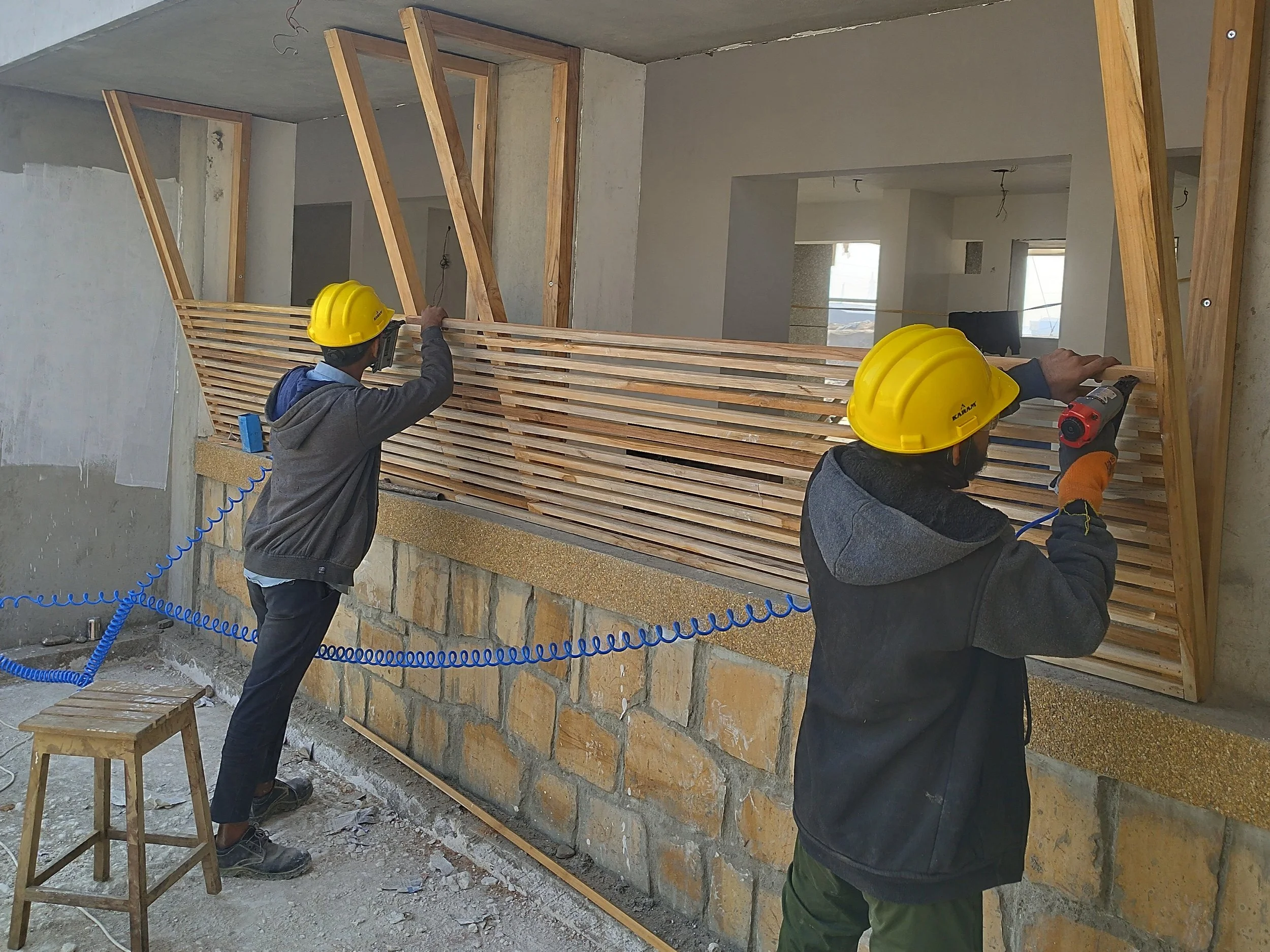 Two construction workers with yellow helmets working on a wooden structure in an unfinished building. One worker is holding a power drill, while the other is adjusting a wooden piece. The background shows a construction site with concrete walls and unfinished interior space.