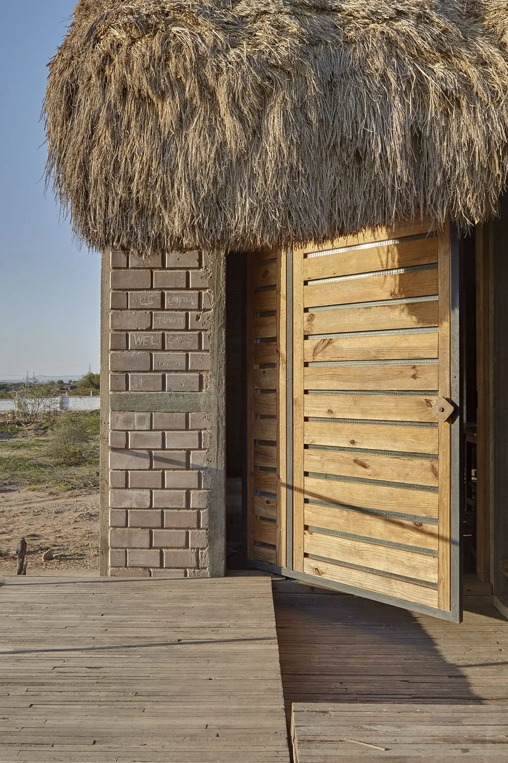 A small building with a thatched roof, brick pillar, and wooden door slightly open, set in a sunny, outdoor environment.