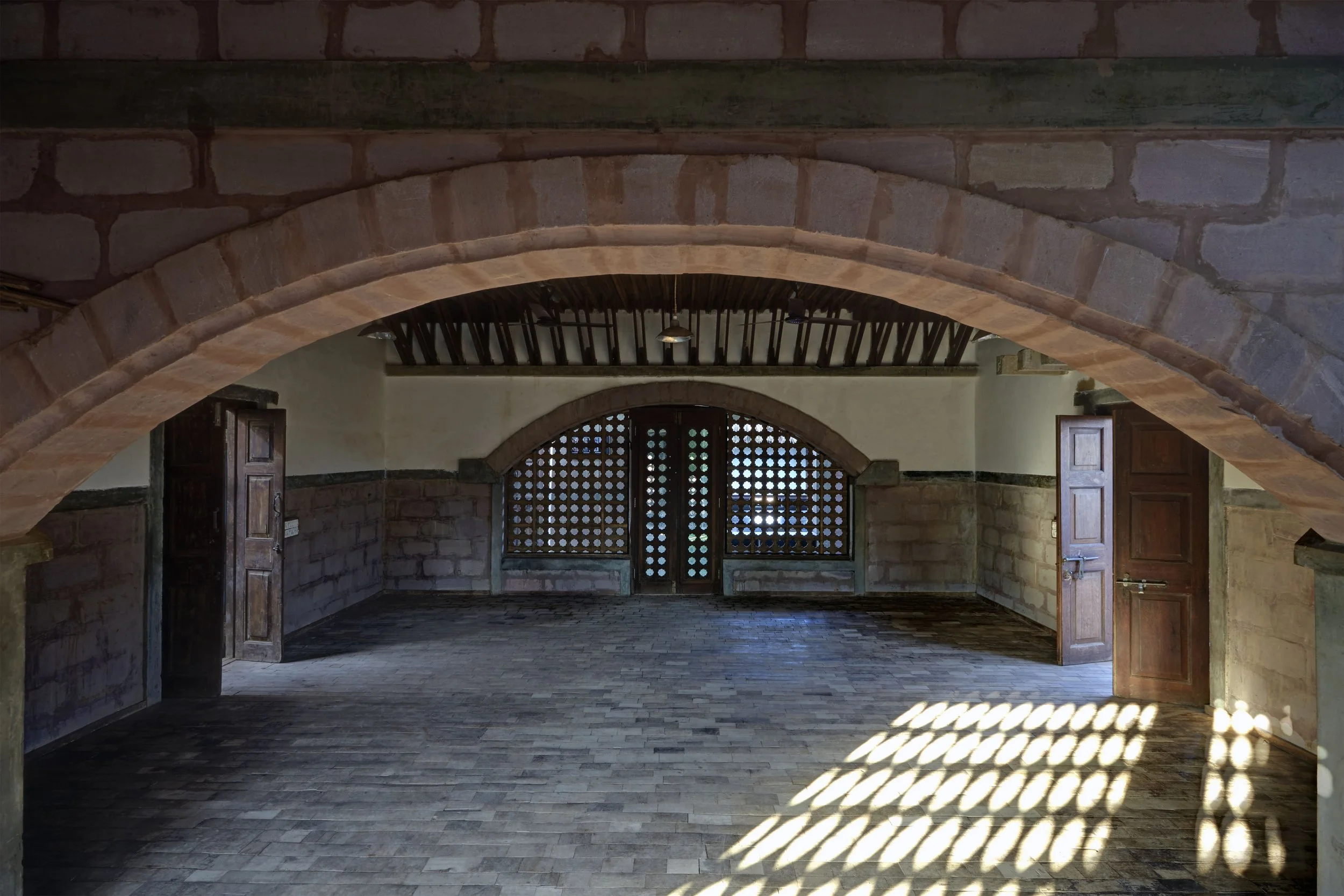 Interior of a stone building with an arched entrance and a door with a lattice window, sunlight casting a pattern on the floor.
