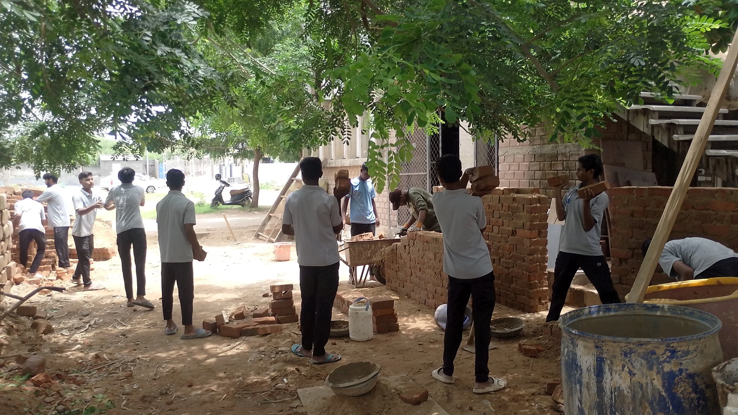 Construction workers building a brick wall under a tree, some carrying bricks, others working with tools