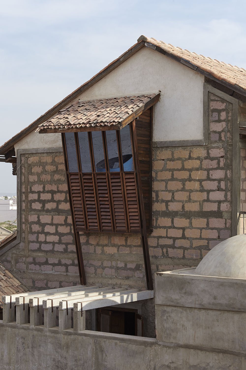 A house with unfinished brick walls, a tiled gable roof, and an unusual window with wooden slats and glass, set against a blue sky.