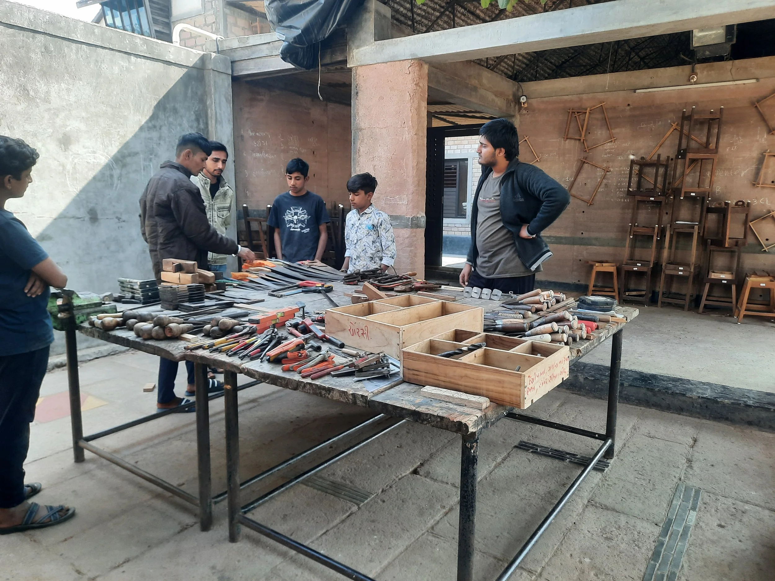 A group of people, including children and adults, gathered around a table displaying various woodworking tools in an outdoor workshop setting.