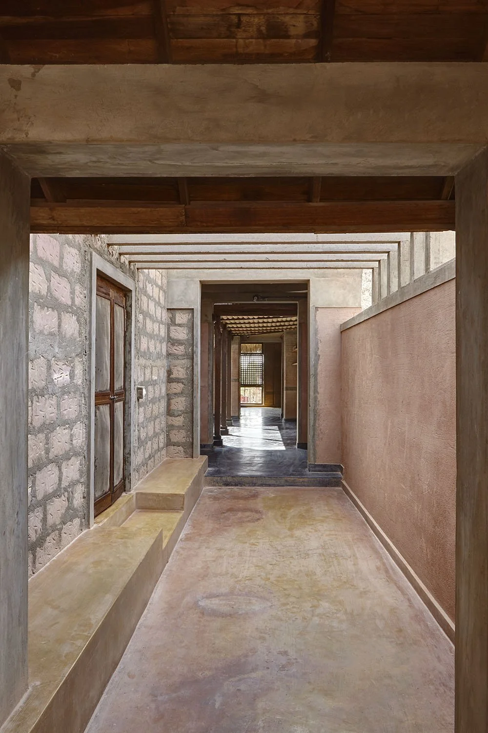 A hallway with stone and pink walls, wooden door, and wooden ceiling beams, leading to aBacklit balcony or outdoor area.