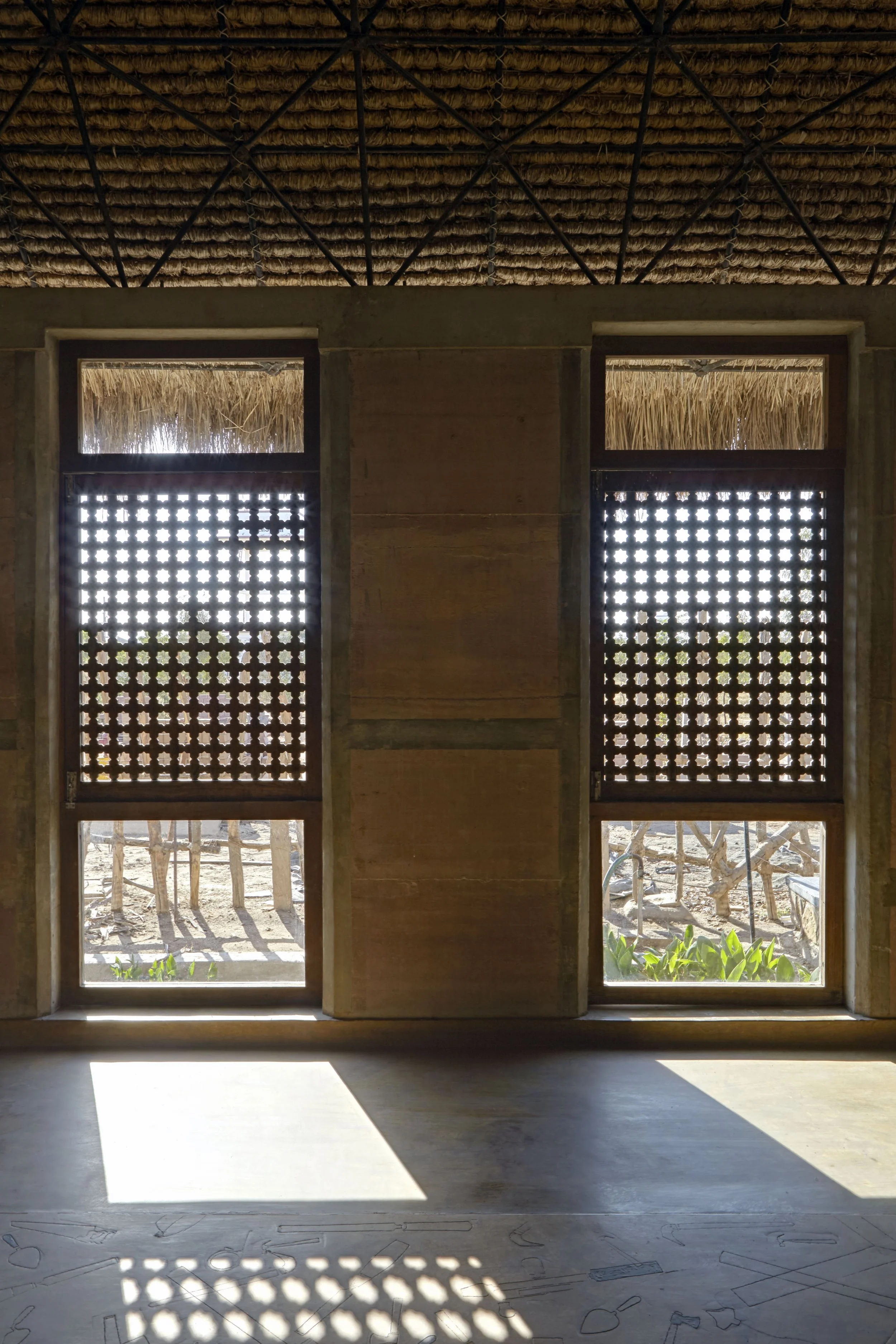 Interior view of a structure with two windows featuring decorative wooden lattices. Sunlight streams through the lattices, casting patterned shadows on the floor. Outside visible through the windows are trees and vegetation.