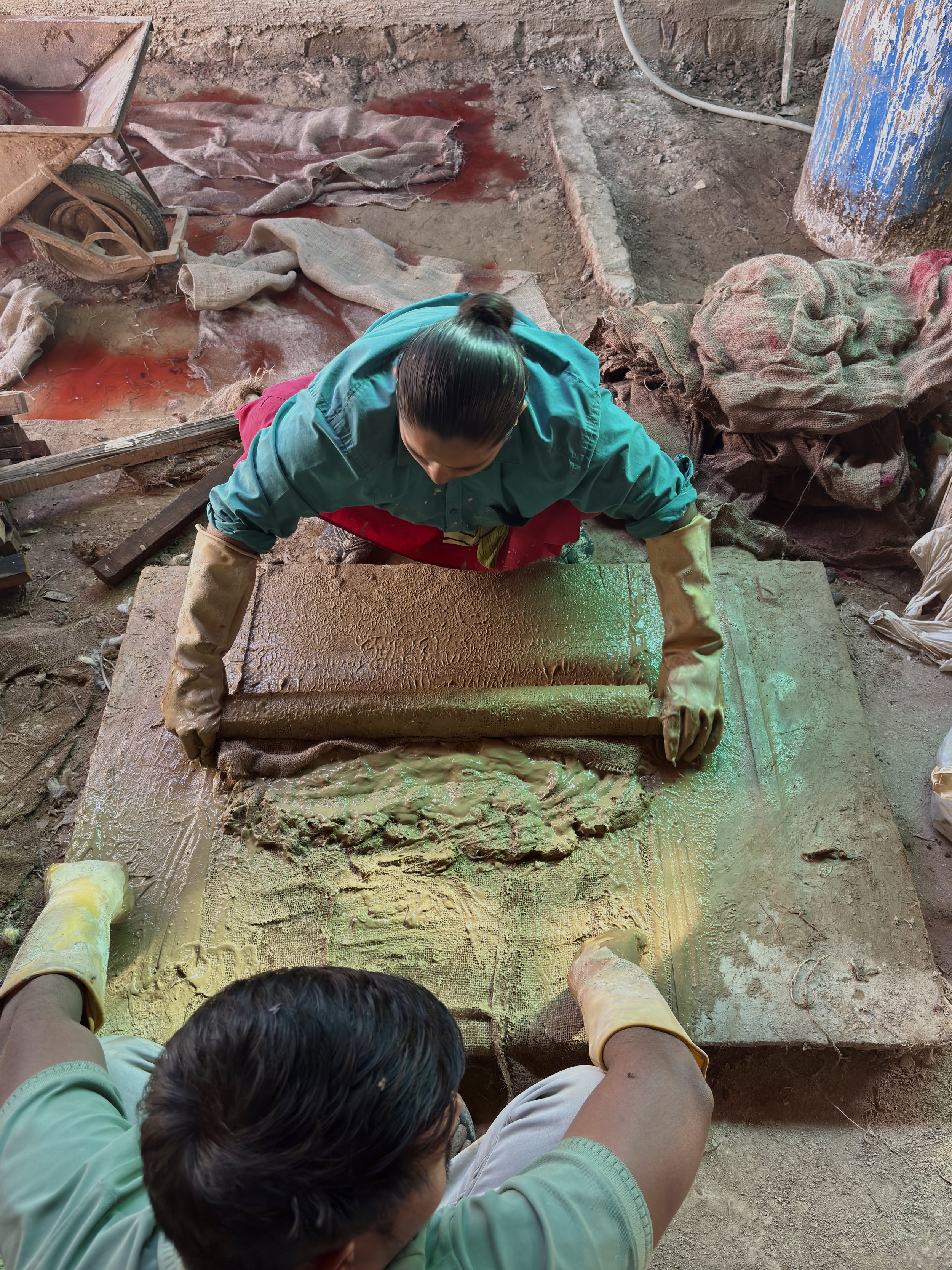 Two people working with clay or mud, using a rolling pin, on a large slab outdoors, surrounded by construction tools and rags.