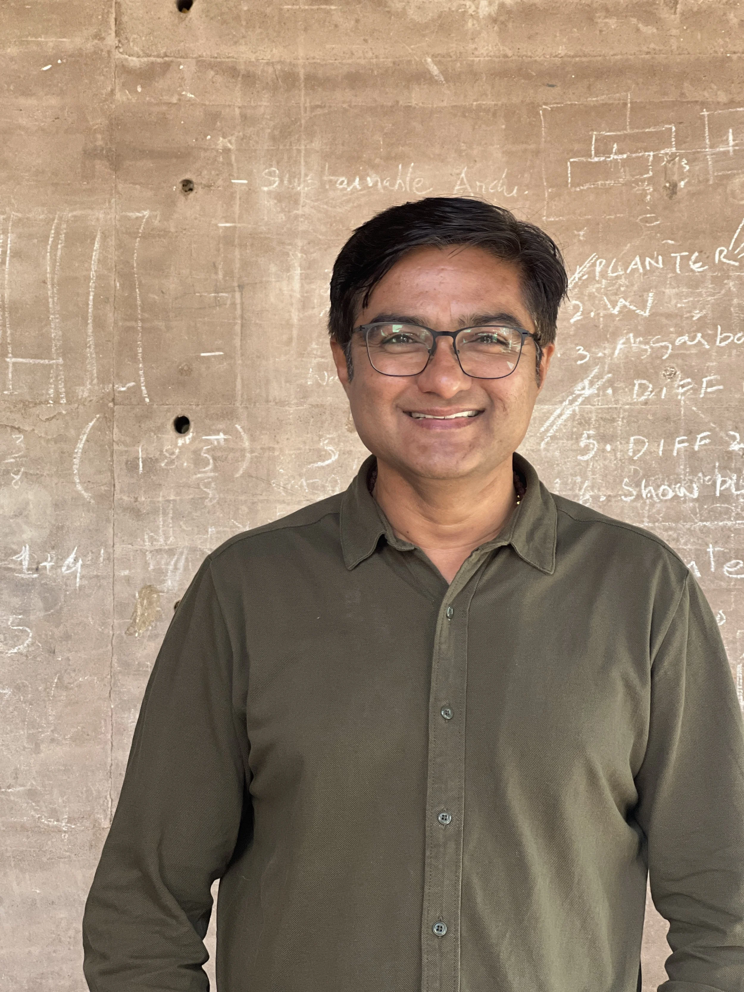 A smiling man with glasses and dark hair, wearing a green button-up shirt, standing in front of a wall with white chalk writing and diagrams.