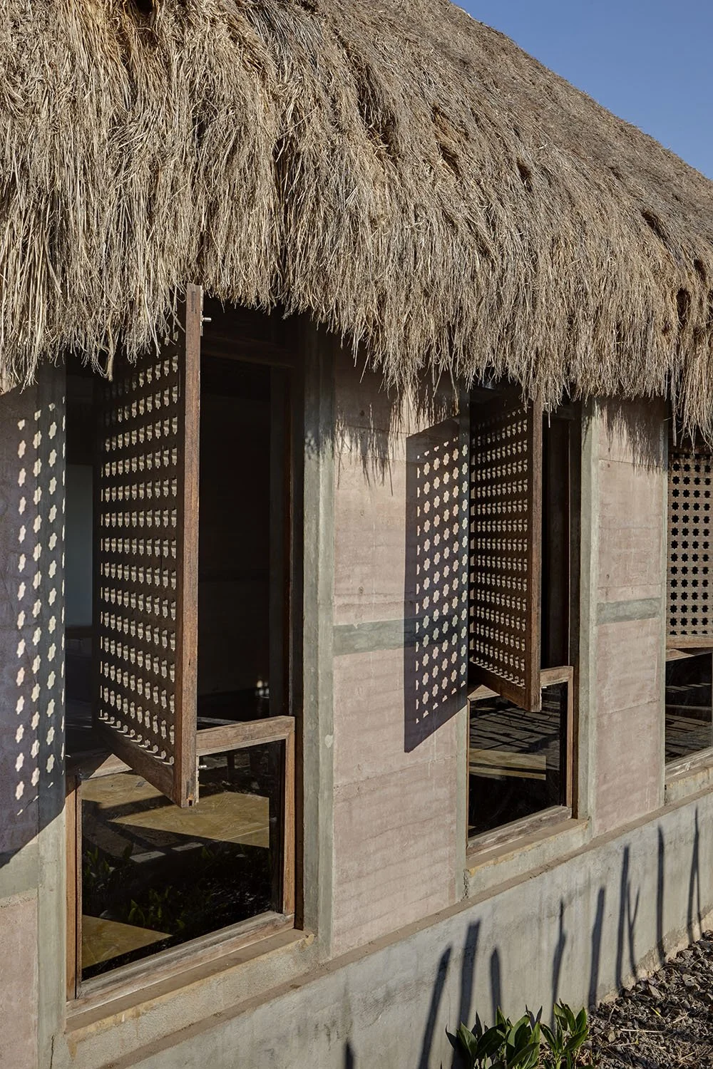 A building with a thatched roof and two open wooden lattice windows casting shadows on the wall.