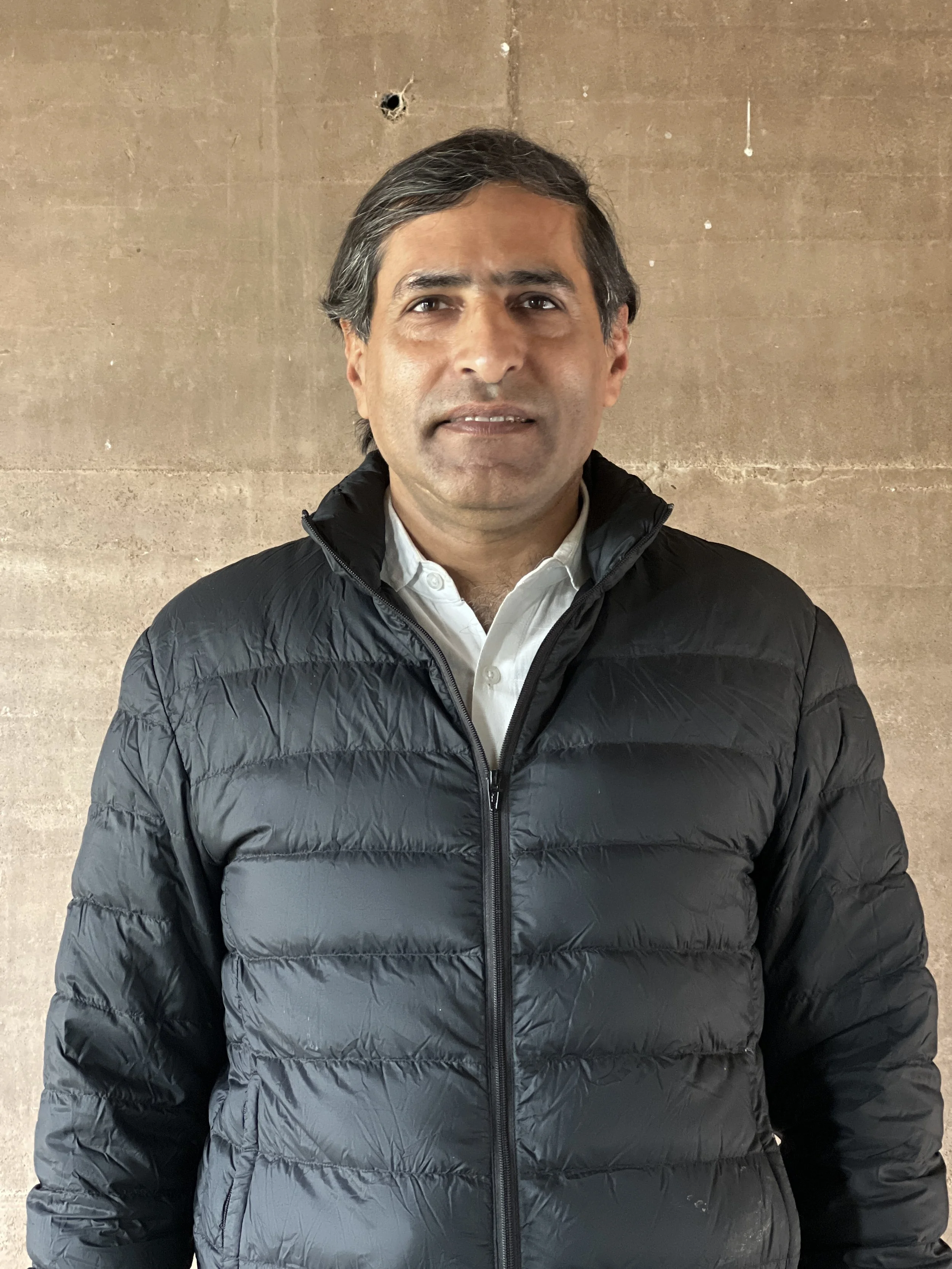 A man with dark hair, wearing a black puffy jacket over a white collared shirt, stands in front of a plain brown wall.