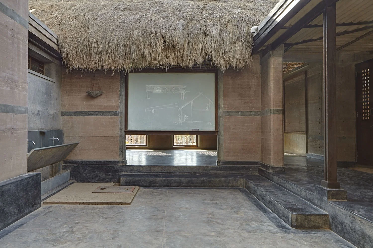 Interior of a rustic building with a thatched roof, concrete walls, and an open space featuring a chalkboard with architectural sketches, a stone sink, and steps leading to another room.