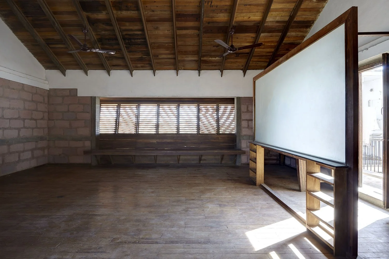 Empty wooden room with brick walls, wooden ceiling with two ceiling fans, bench along brick wall, and a large whiteboard on a wooden stand near an open door.