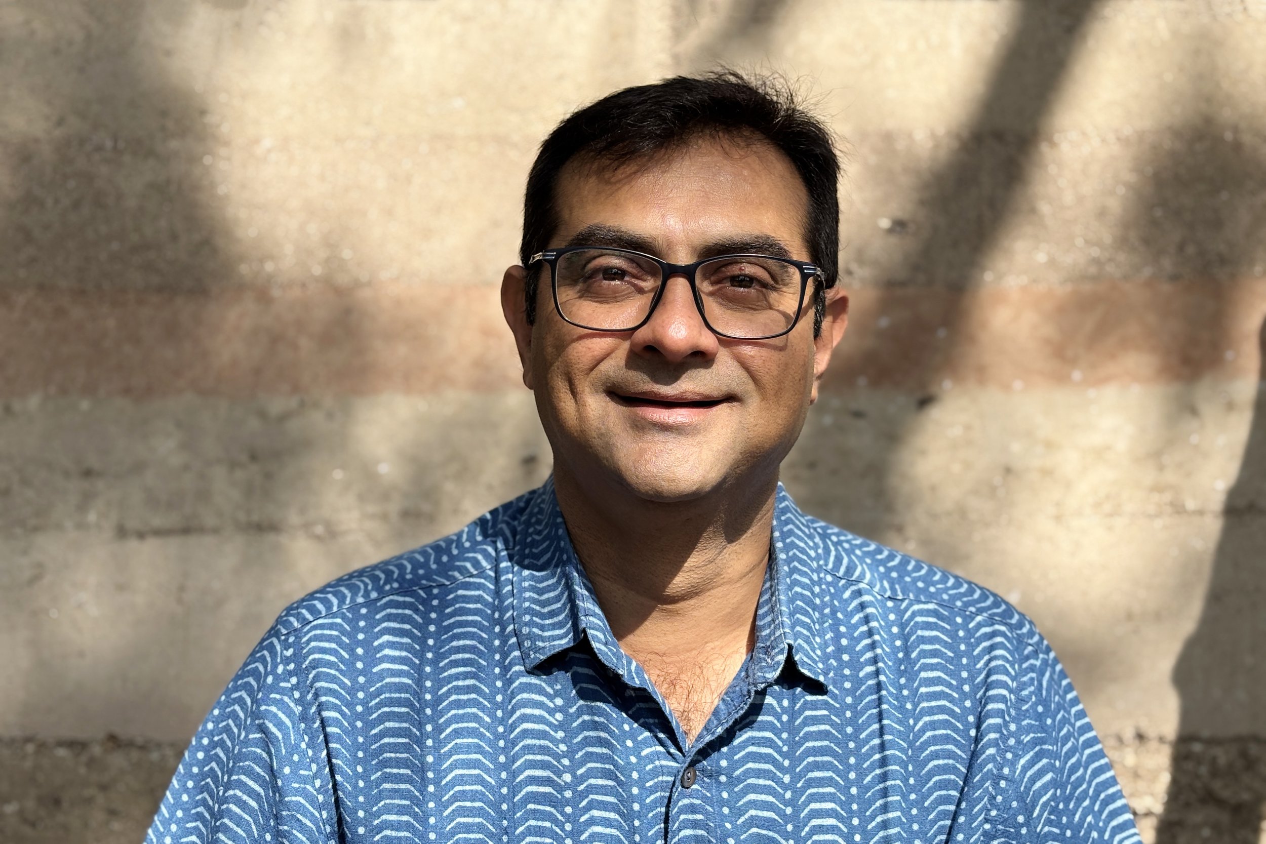 Man with glasses wearing a blue patterned shirt smiling outdoors