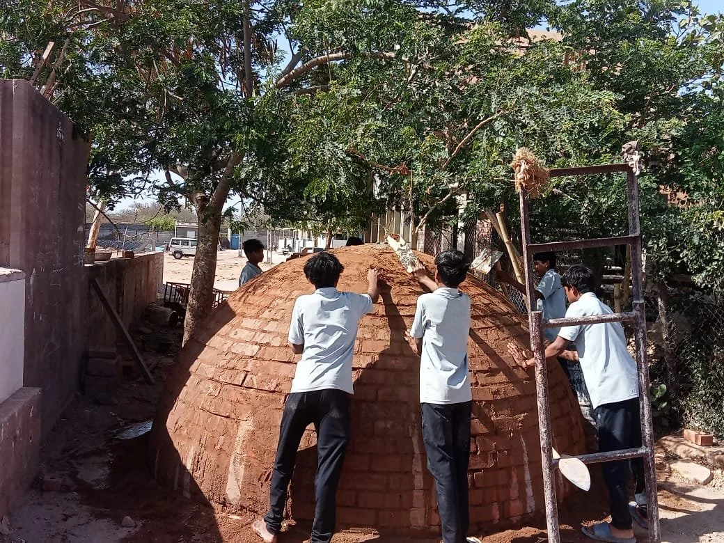 Group of students working on building a large brick structure outdoors with trees and a fence in the background.