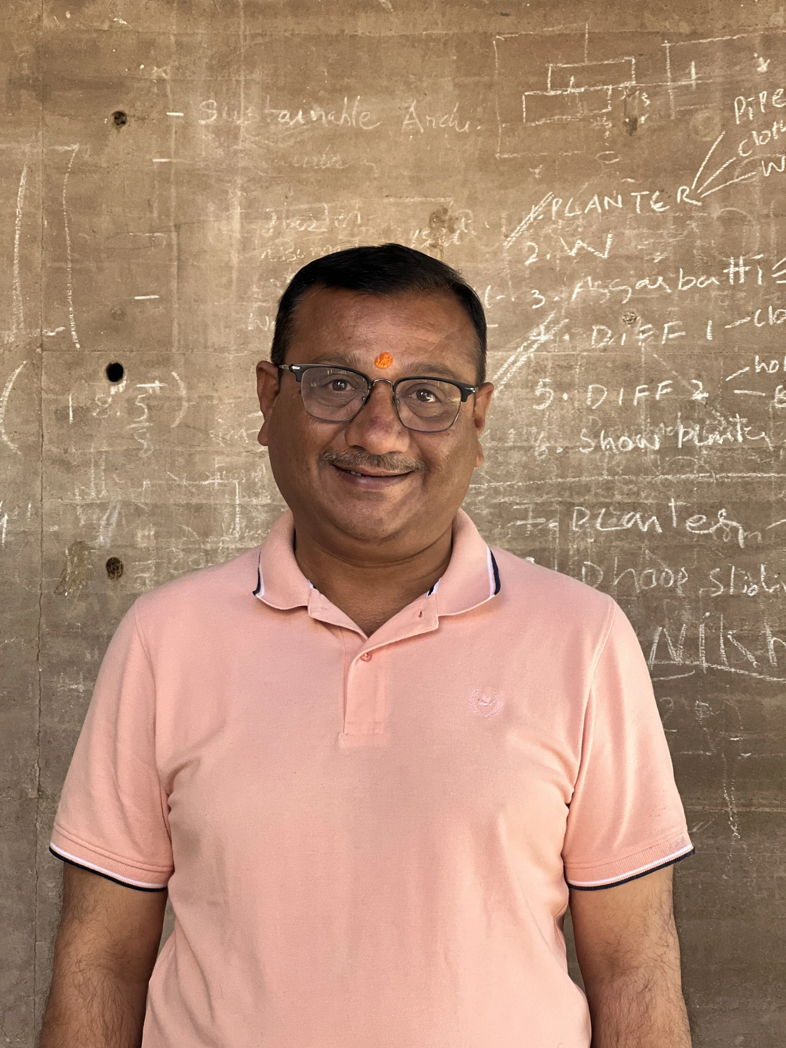 A smiling man wearing glasses and a pink polo shirt standing in front of a chalkboard with notes and diagrams
