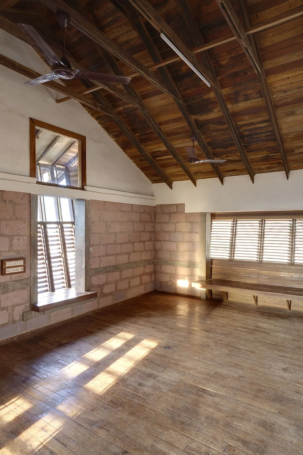 An empty room with a wooden floor, brick and plaster walls, a wooden ceiling with exposed beams, and ceiling fans. There are two windows with wooden blinds, one small square window and one larger window with a bench underneath.