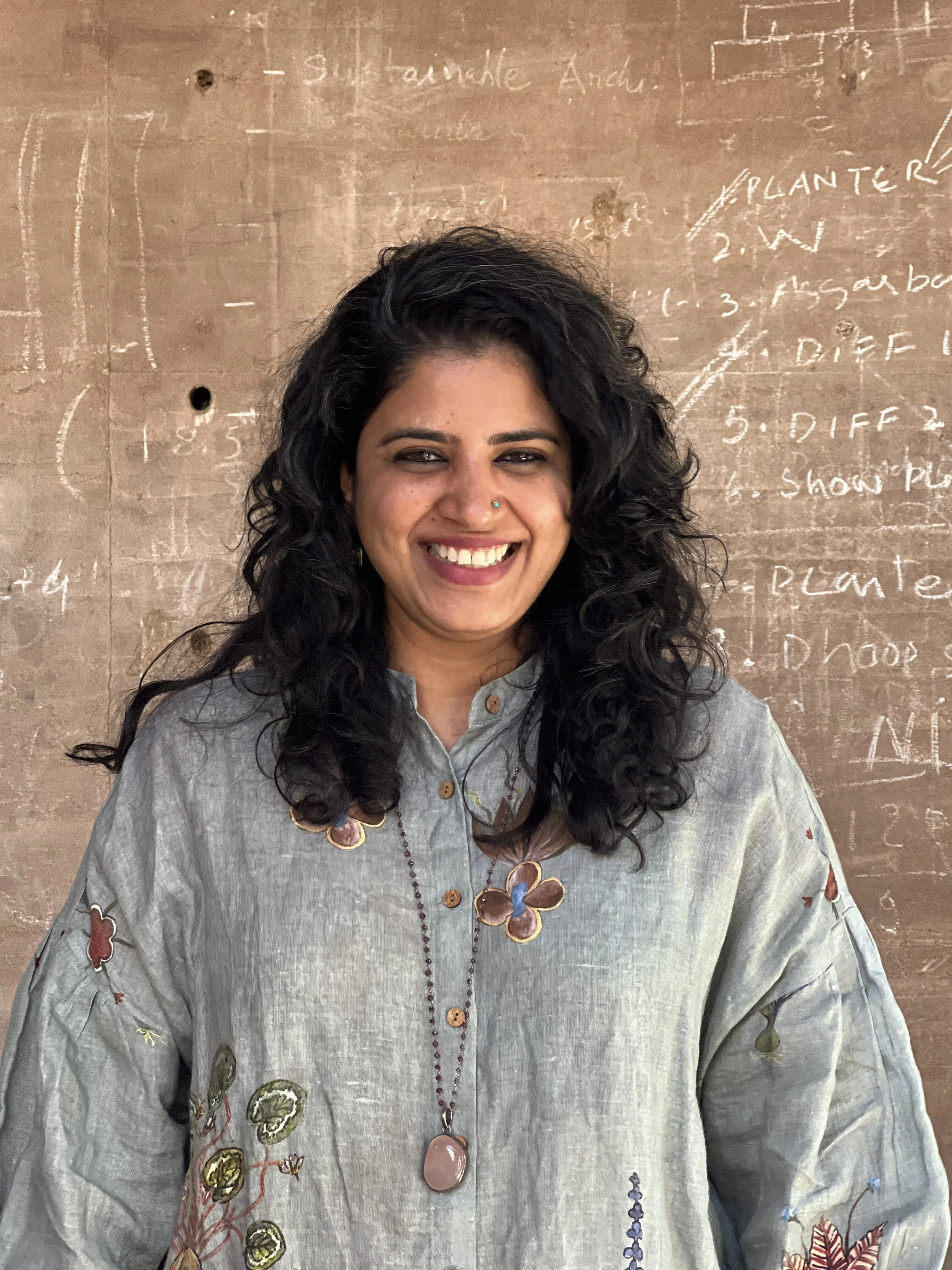 A woman with long, curly black hair and a nose piercing smiling, wearing a light gray embroidered shirt and a beaded necklace, standing in front of a brown wall with handwritten notes and diagrams.