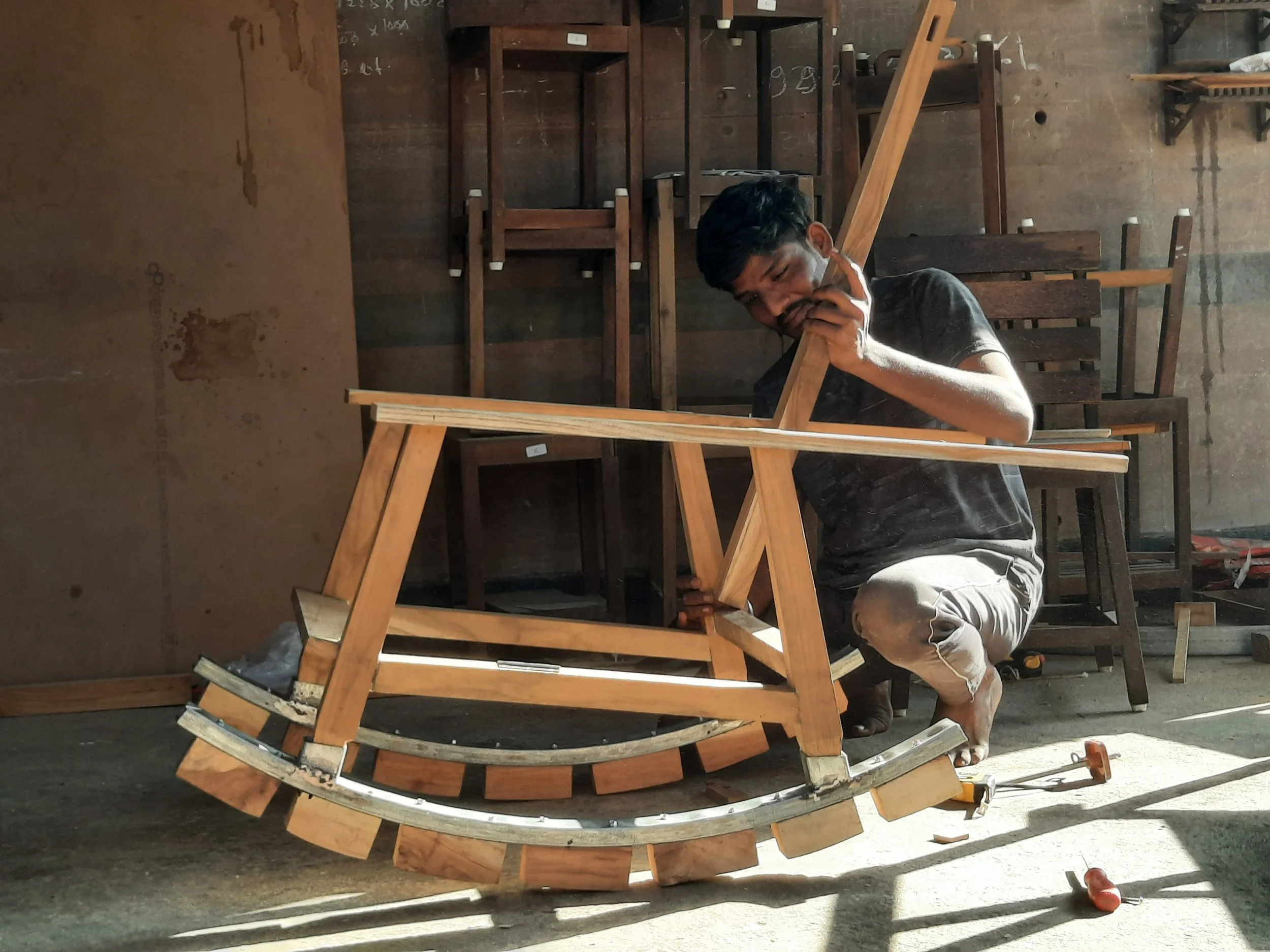 A man is working on a wooden rocking chair in a workshop, surrounded by other wooden chairs and tools, with sunlight casting shadows on the floor.