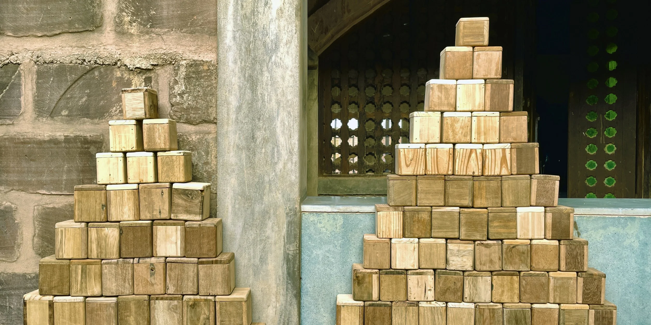 Two stacks of wooden blocks arranged in pyramid shapes, positioned against an old stone and concrete wall with a decorative wooden window in the background.