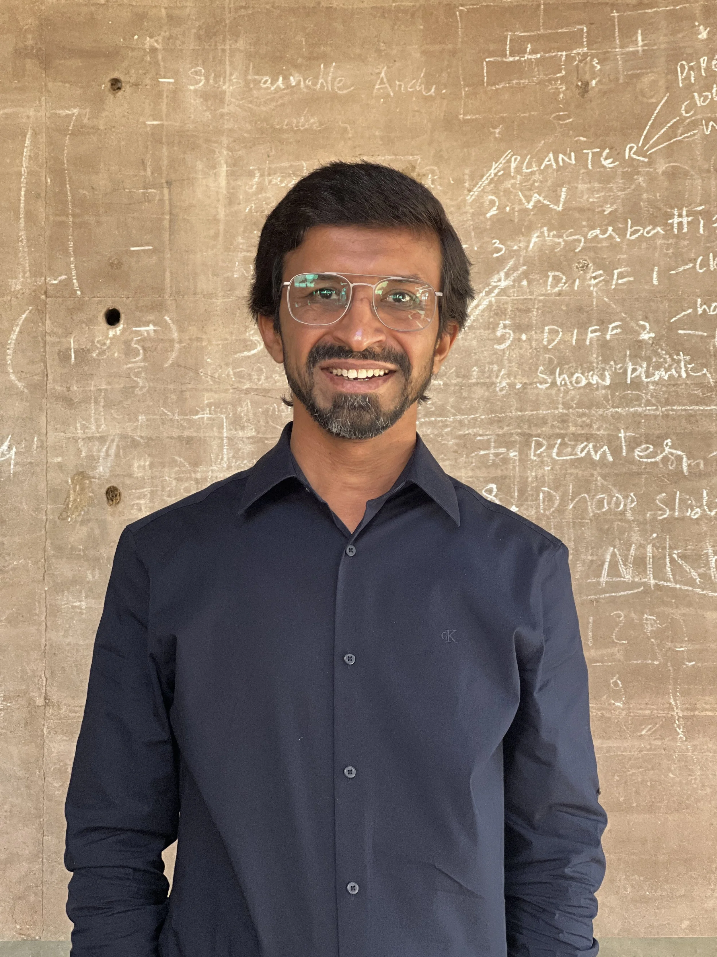 A man with glasses and a beard, wearing a dark blue button-up shirt, smiling in front of a wall with chalk writing and diagrams.