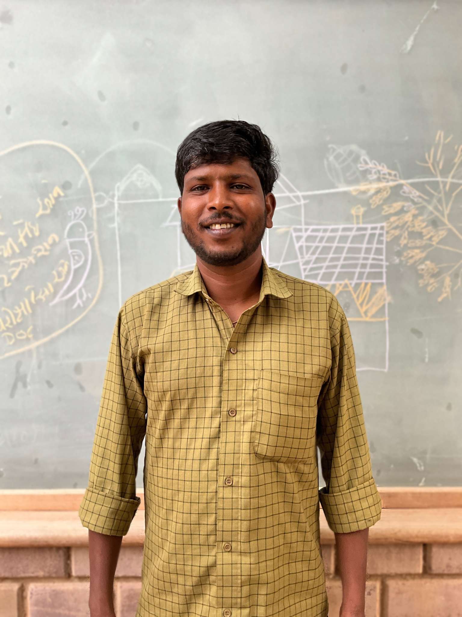 A smiling man with short dark hair and a beard standing in front of a wall with writing on it, wearing a beige polo shirt.