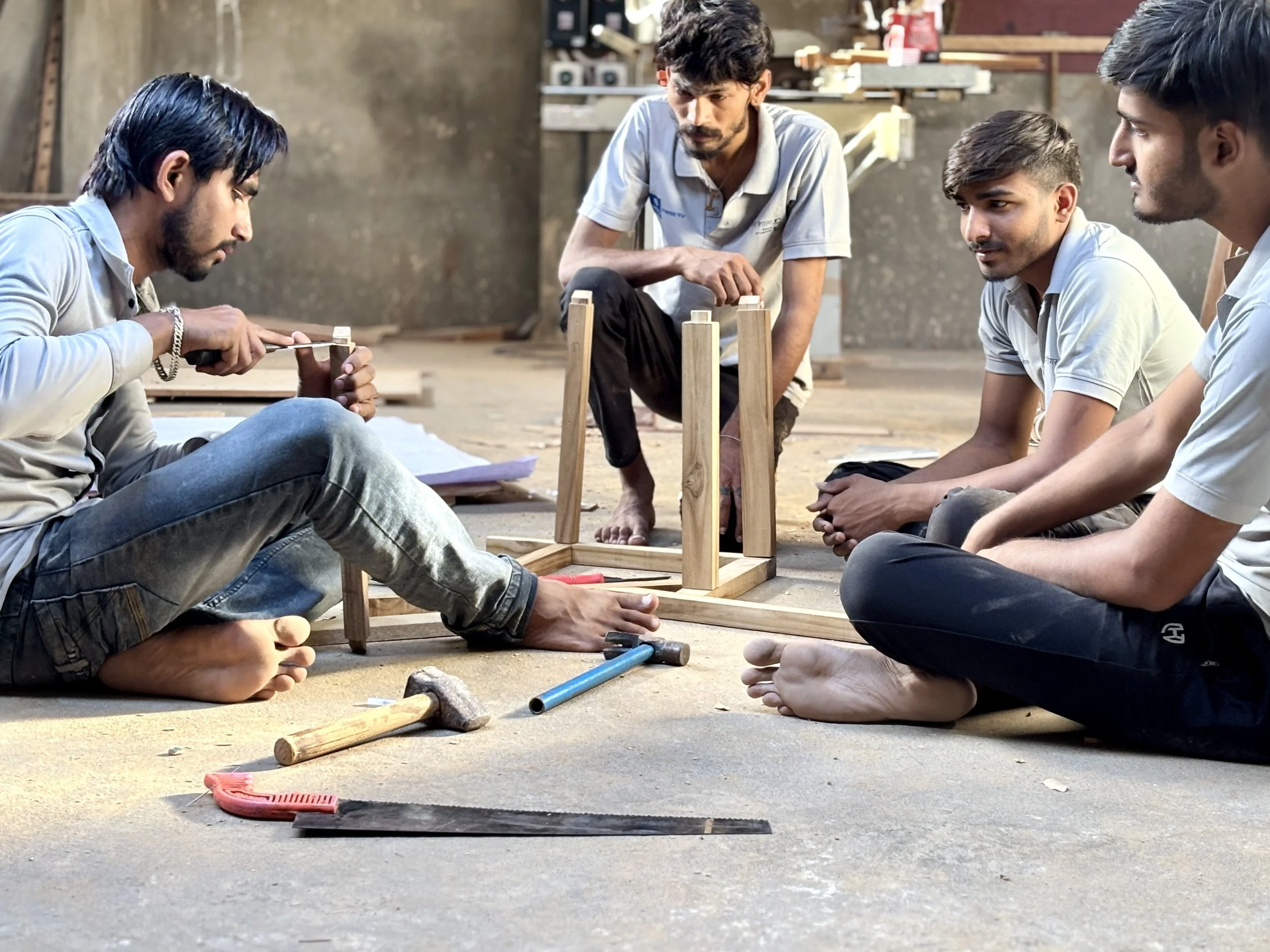 Four young men working on assembling a wooden stool in a workshop, sitting on the floor surrounded by tools.
