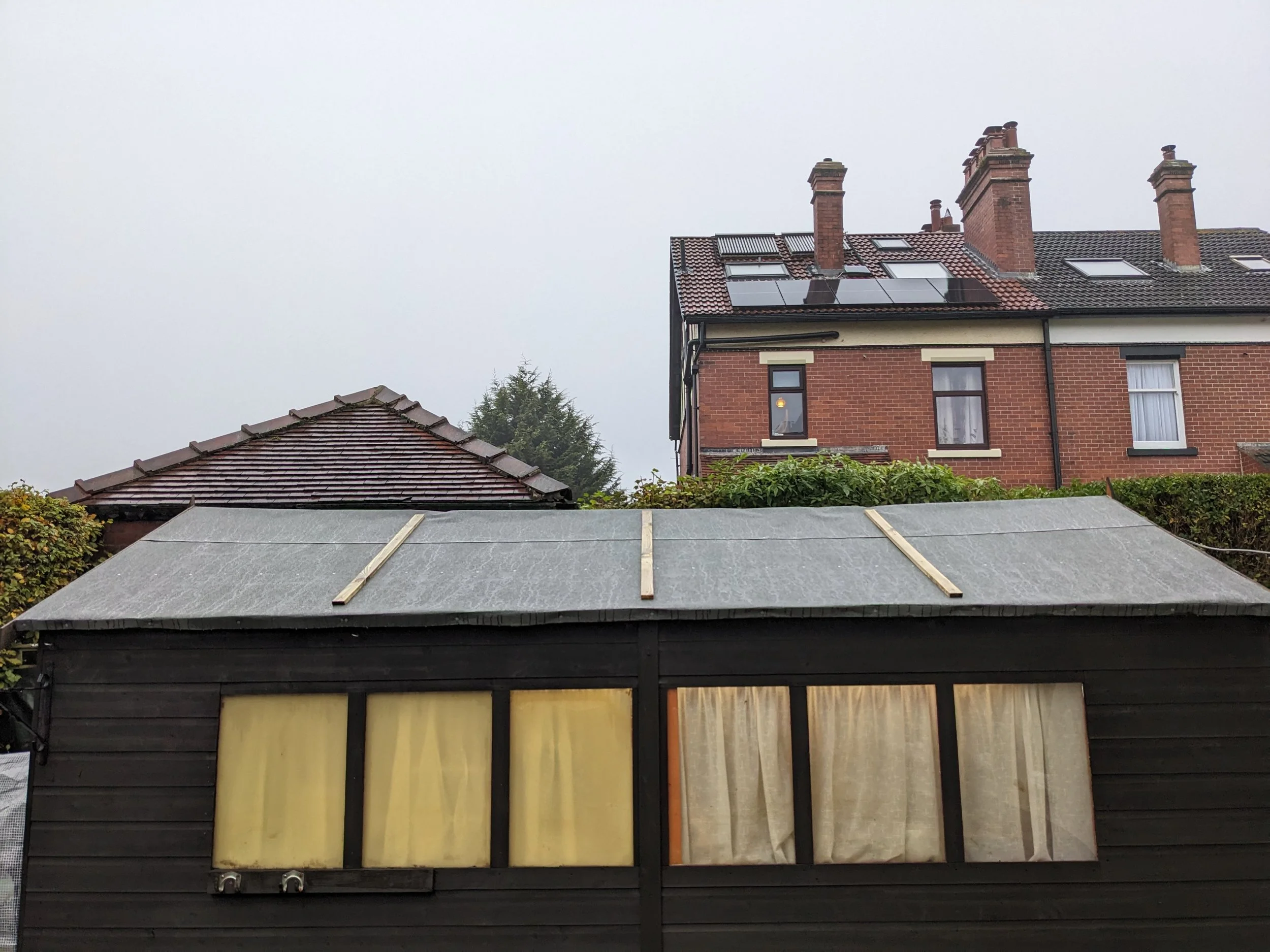 A black wooden building with yellow curtains and a slanted roof covered with a grey tarp, with an older brick house in the background on a cloudy day.