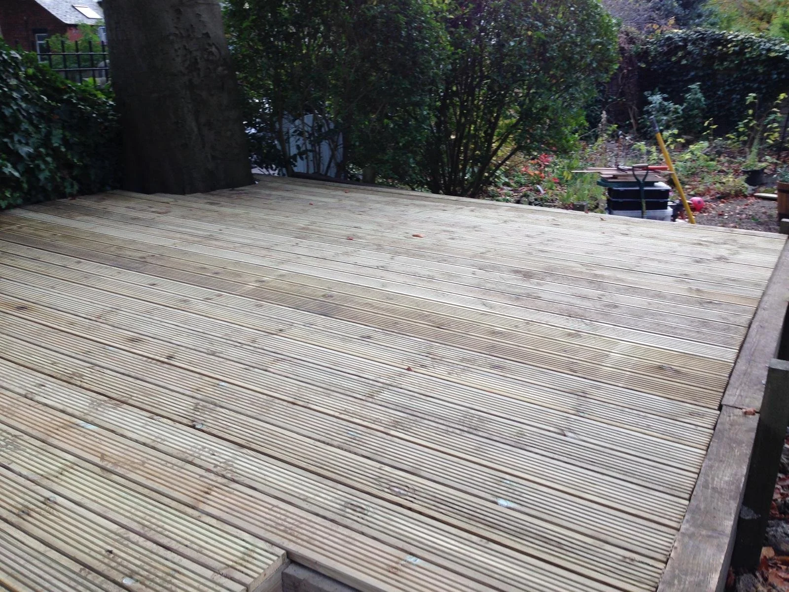 A newly constructed wooden deck with horizontal planks, surrounded by a garden with bushes and trees.