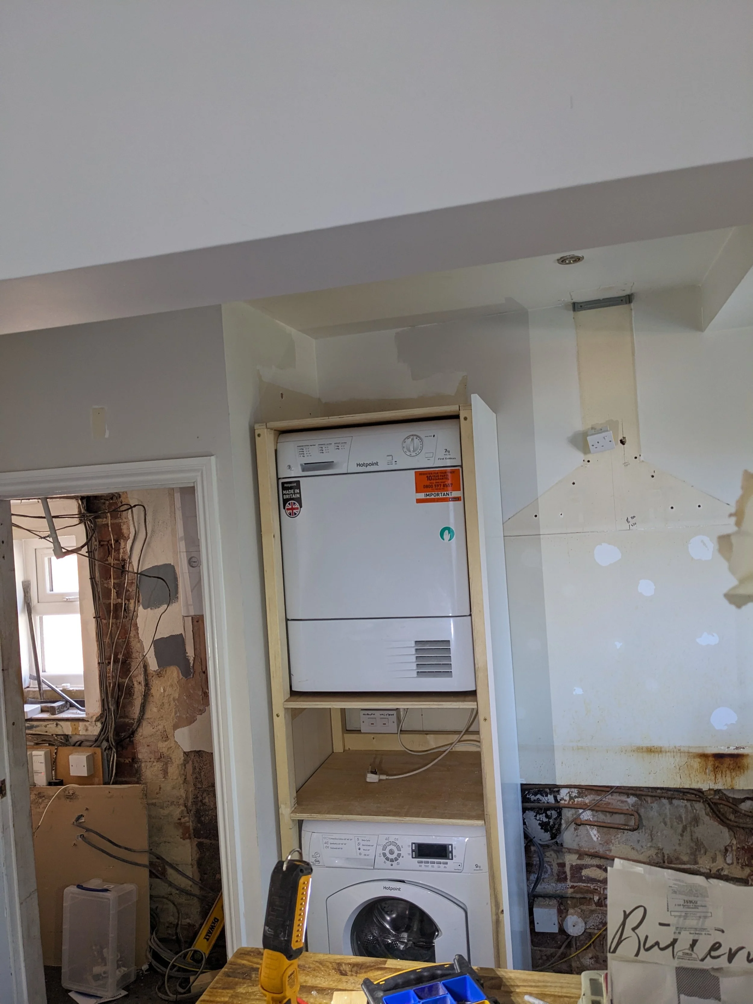 A partially installed laundry area with a boiler, washing machine, and exposed wiring and pipes in a room under renovation.