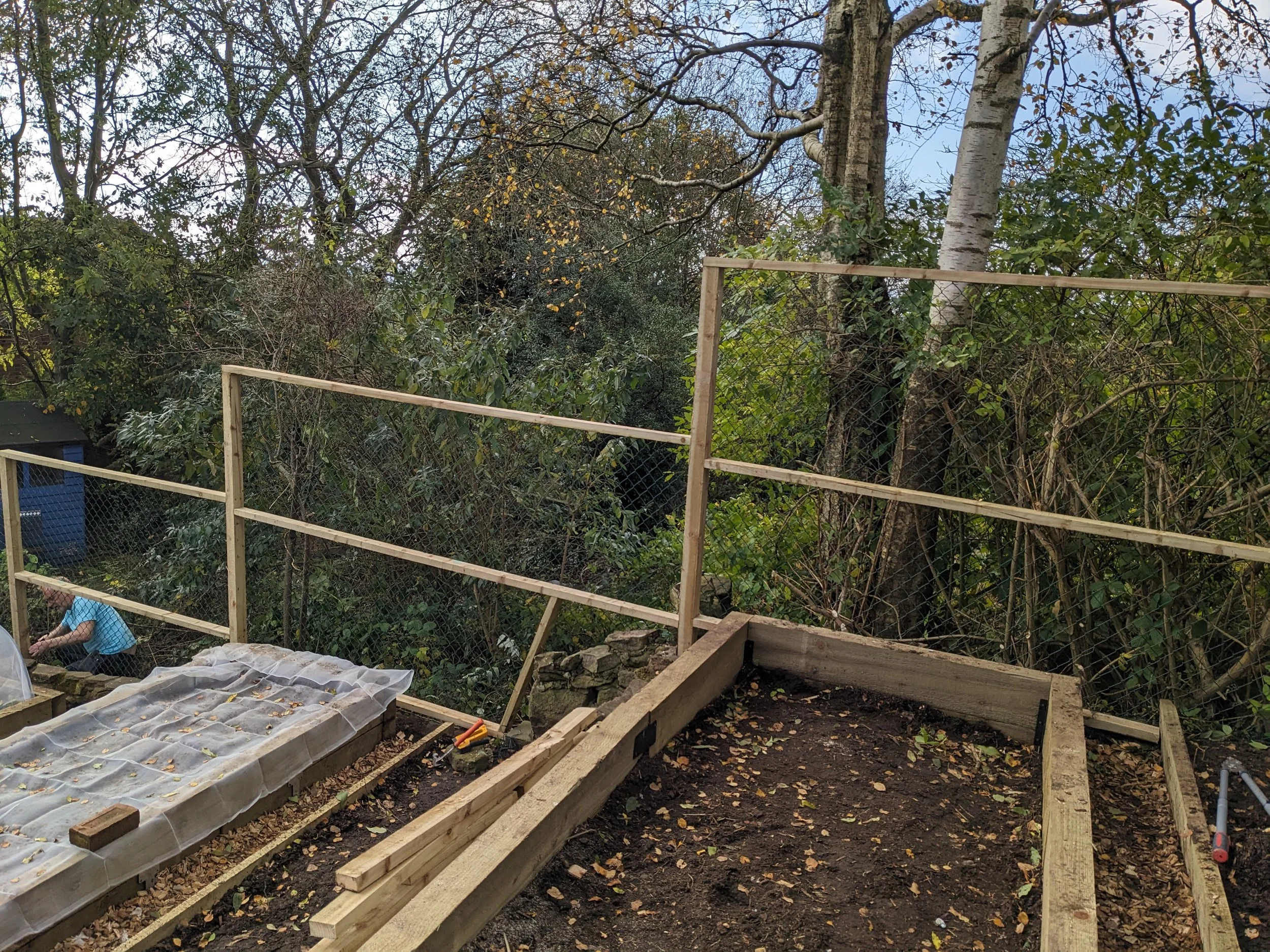 A garden construction site with wooden frames and fencing, surrounded by trees and foliage, during autumn.