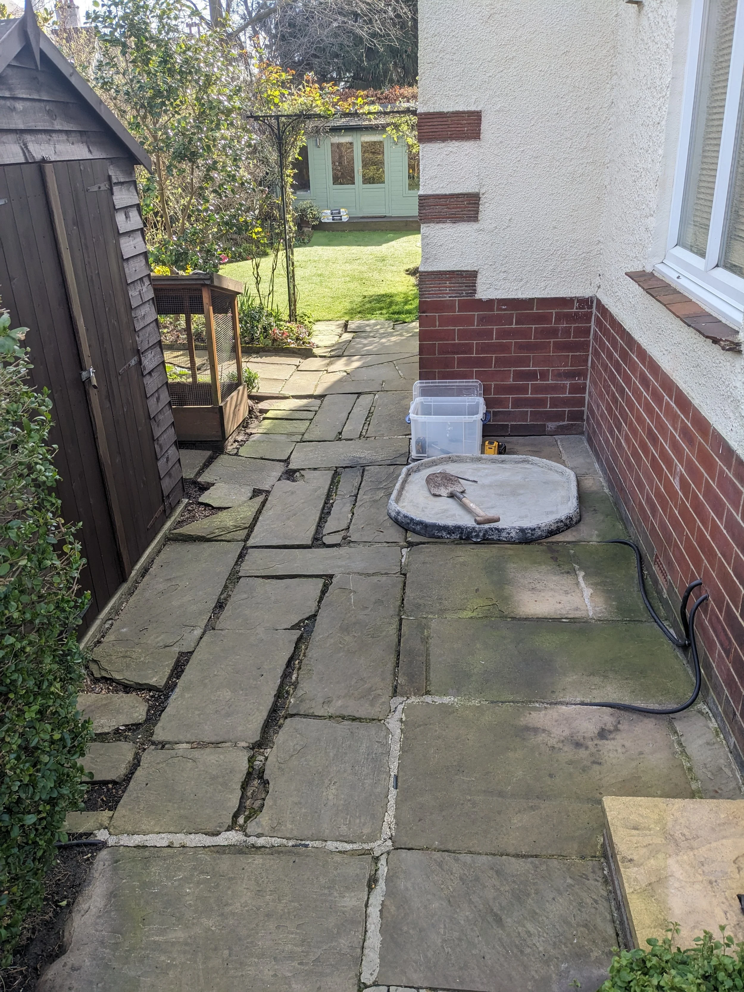 A narrow backyard patio with large stone slabs leading to a grassy lawn. A small shed on the left, a plastic container and a trowel on a flat stone in the center, and a house wall with a window on the right.