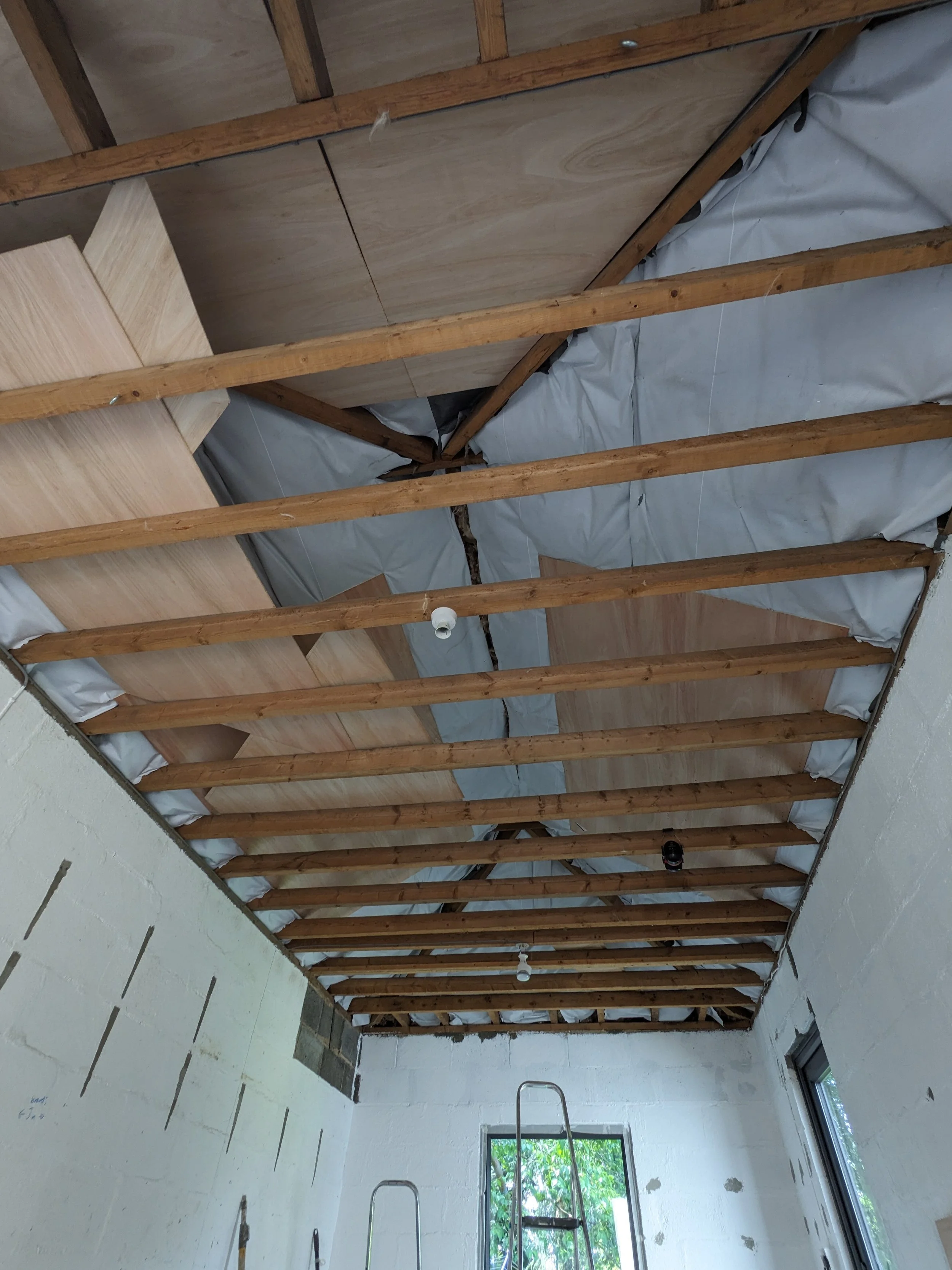 Interior of a room under construction with exposed wooden ceiling joists, insulation, and unfinished walls. Two ladders are positioned inside, and there is a window showing greenery outside.