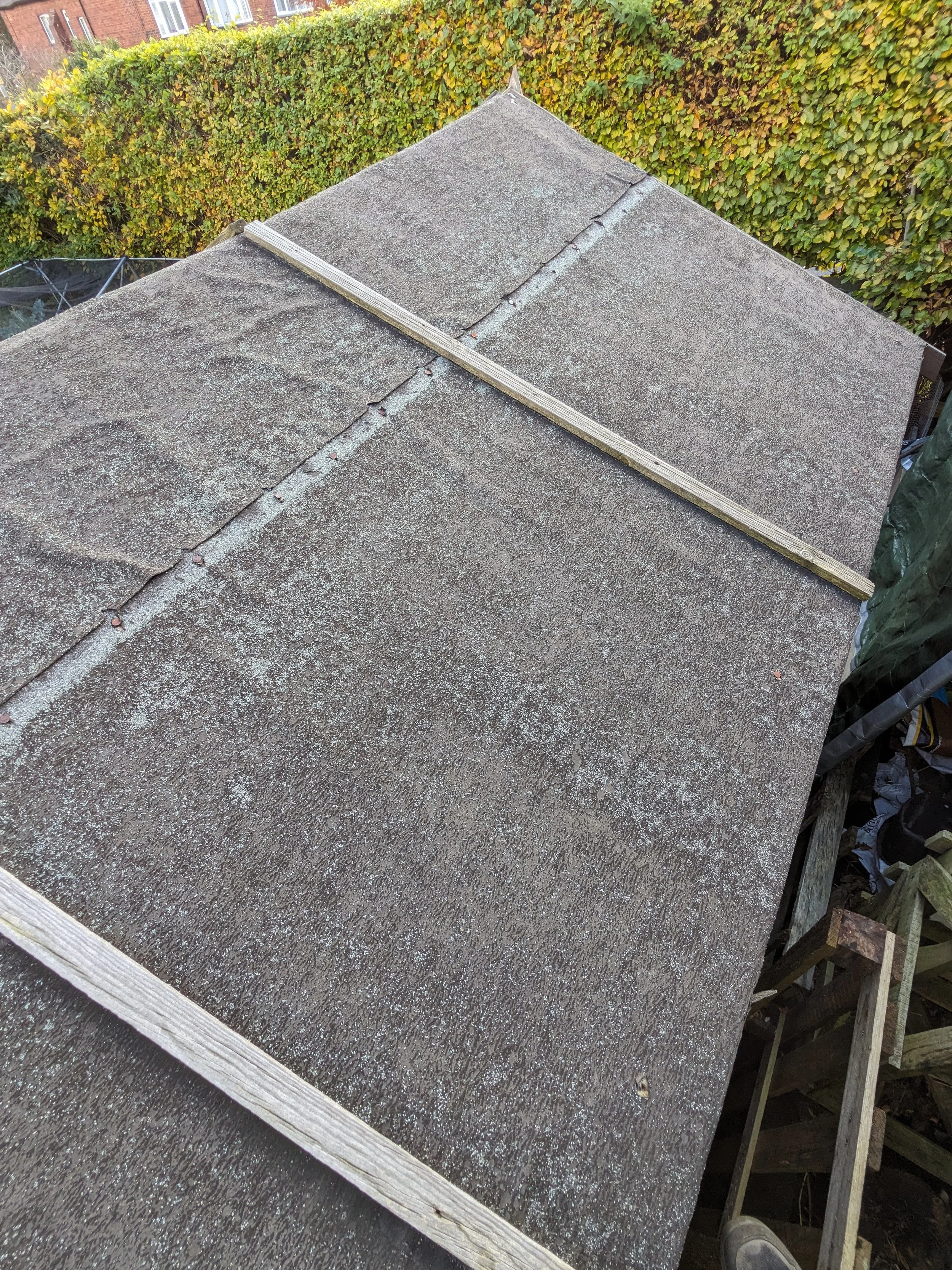 A bird's-eye view of a sloped rooftop with weathered shingles, with a wooden plank placed across part of it, and green foliage in the background.
