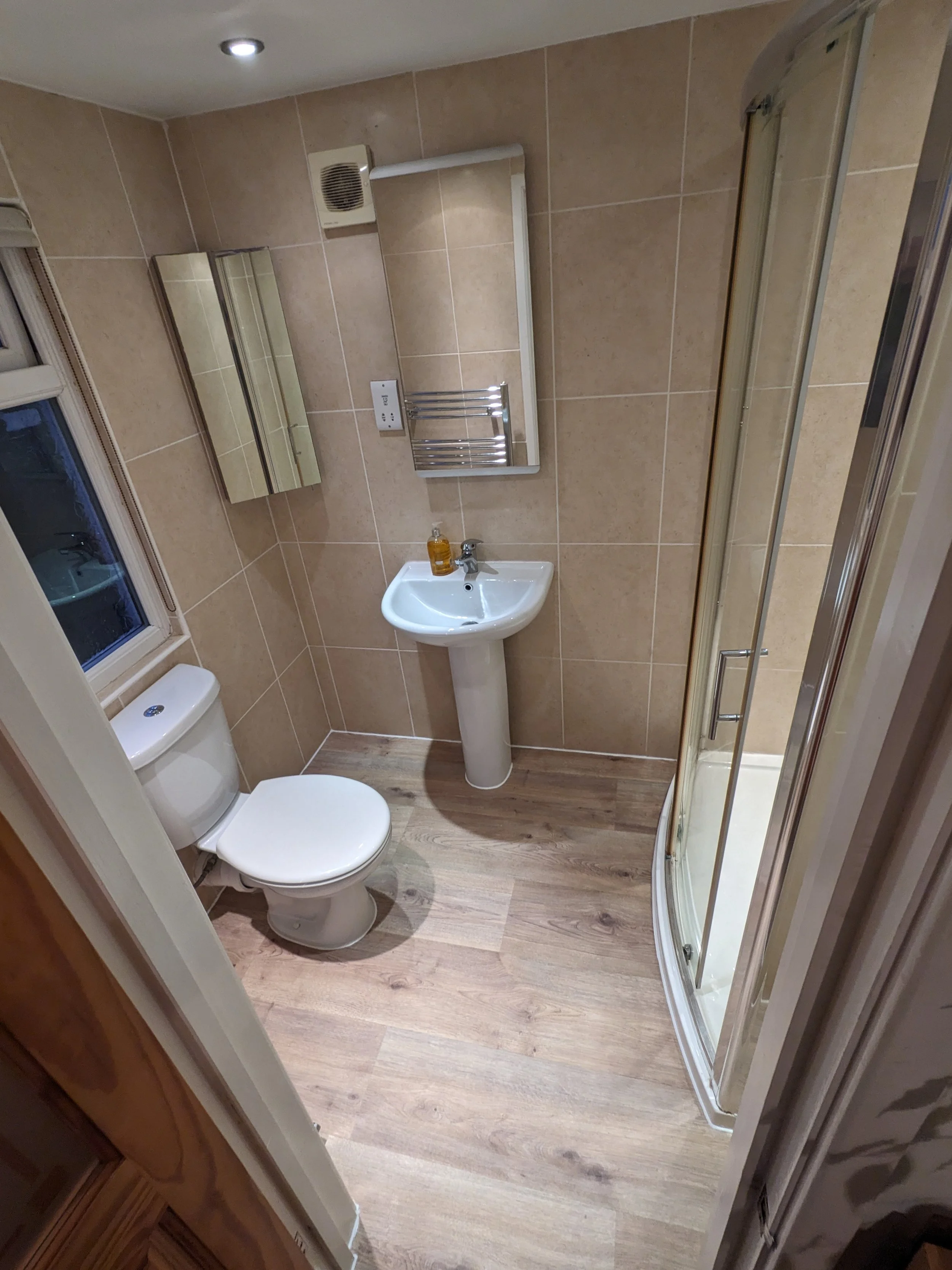 Small bathroom with beige tiled walls, a toilet, a small pedestal sink with hand soap, a rectangular mirror, a wall-mounted towel rack, and a curved shower stall with a glass door. There is a window with blinds and a ceiling light fixture.