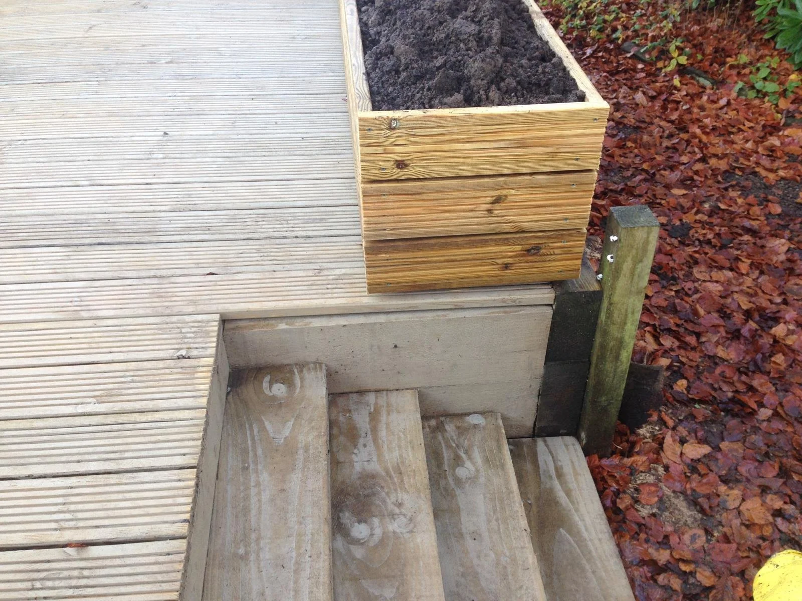 A section of an outdoor wooden deck with stairs, a wooden planter box filled with dark soil, and fallen autumn leaves surrounding the area.