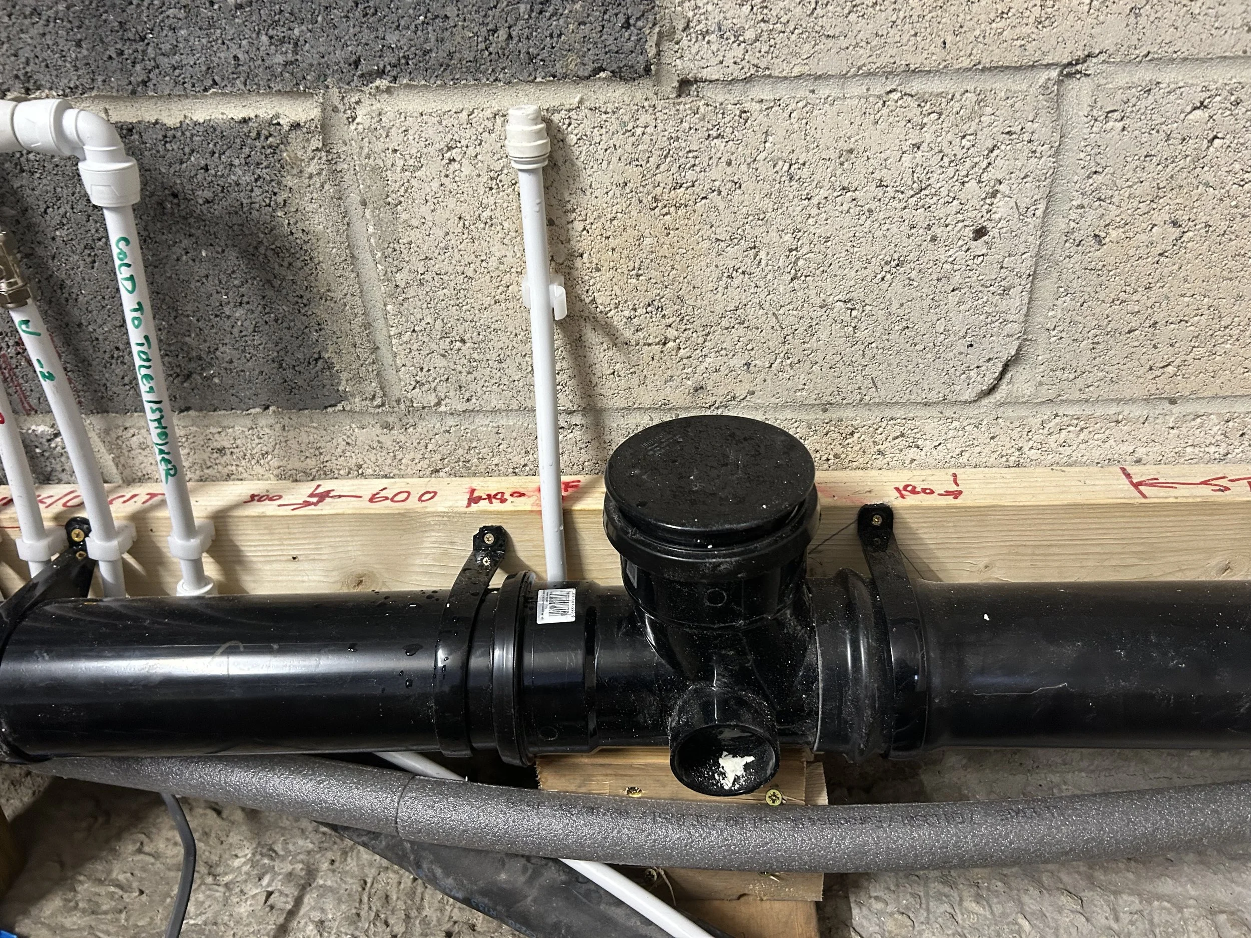 Close-up of plumbing pipes and fittings against a concrete wall, with handwritten measurements on a wooden board.