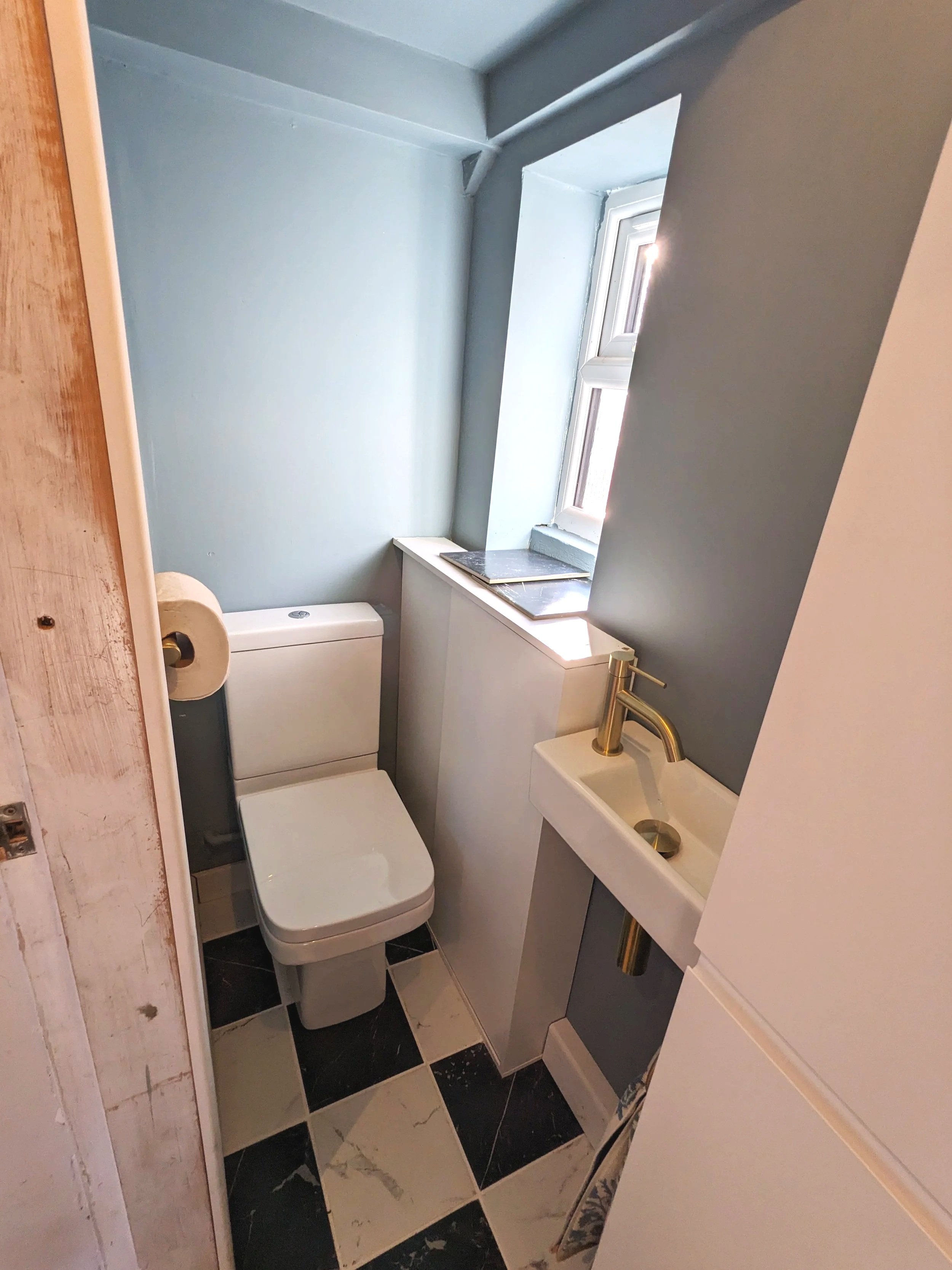Small bathroom with a toilet, small sink with gold fixtures, window letting in natural light, and black and white checkered floor tiles.