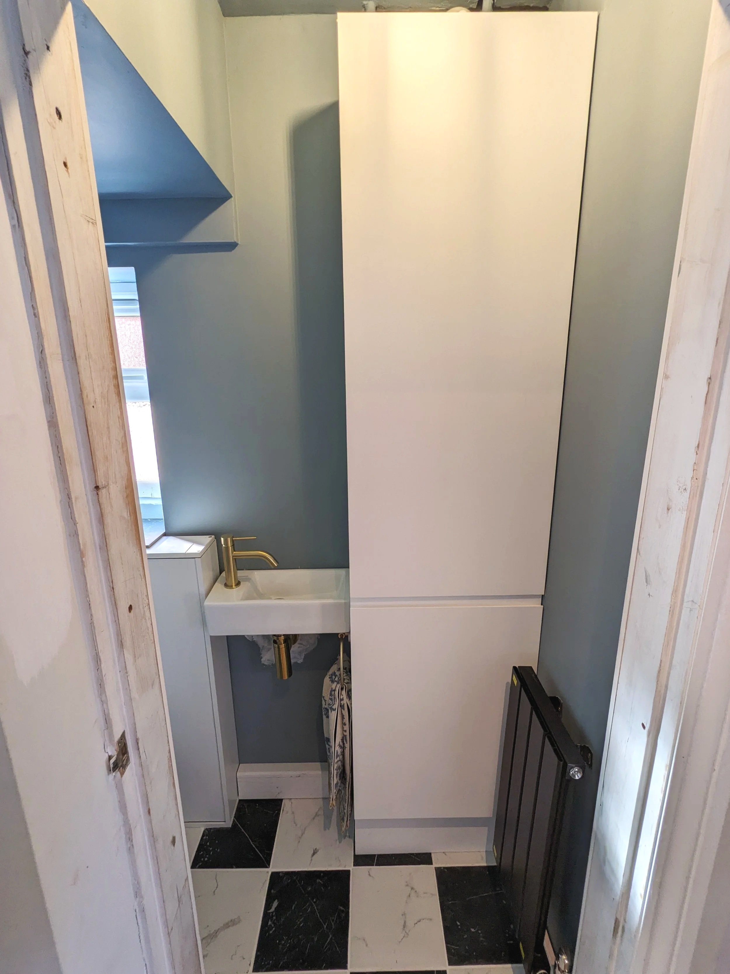 Small laundry or utility room with a white mini sink, gold faucet, a tall white cabinet, a black radiator, a small window with blinds, blue painted walls, and black and white checkered floor tiles.