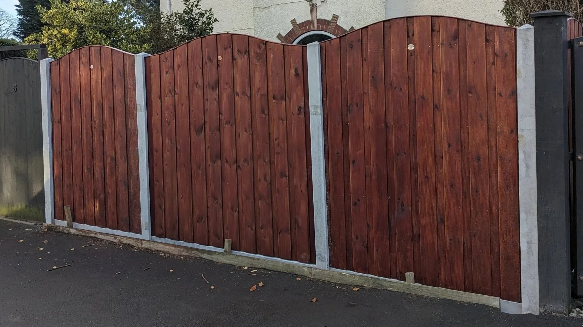 Wooden privacy fence with a curved top, supported by concrete posts, along a paved street in front of a house with a brick arch window and surrounding greenery.