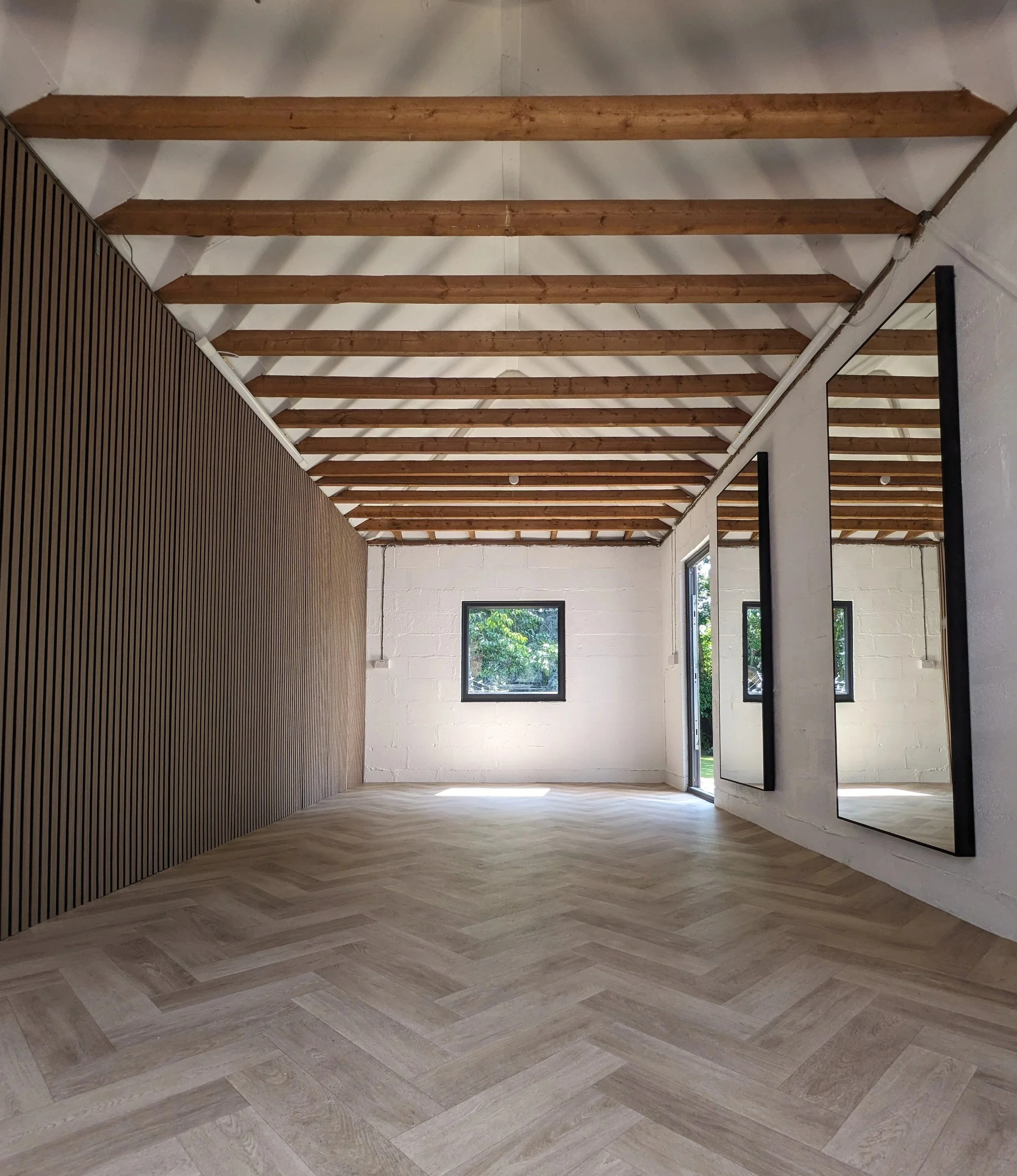 Empty room with light wooden herringbone floor, white brick walls, exposed wooden ceiling beams, a window, and large mirror panels on one wall.