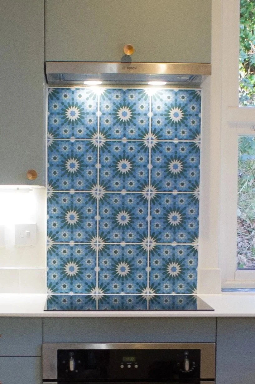 Kitchen stove with a colorful blue and white tiled backsplash, a range hood with lights turned on, and a window showing greenery outside.