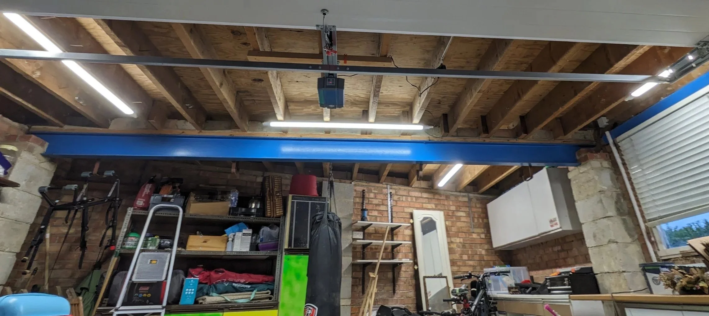 Garage interior with brick walls and exposed wooden ceiling, cluttered with shelves, tools, bicycle, and storage items, featuring a blue garage door track and a garage door opener.