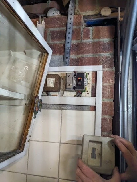 Person holding a beige electrical switch cover near an electrical outlet and circuit breaker on a wall with brick and tiled sections.